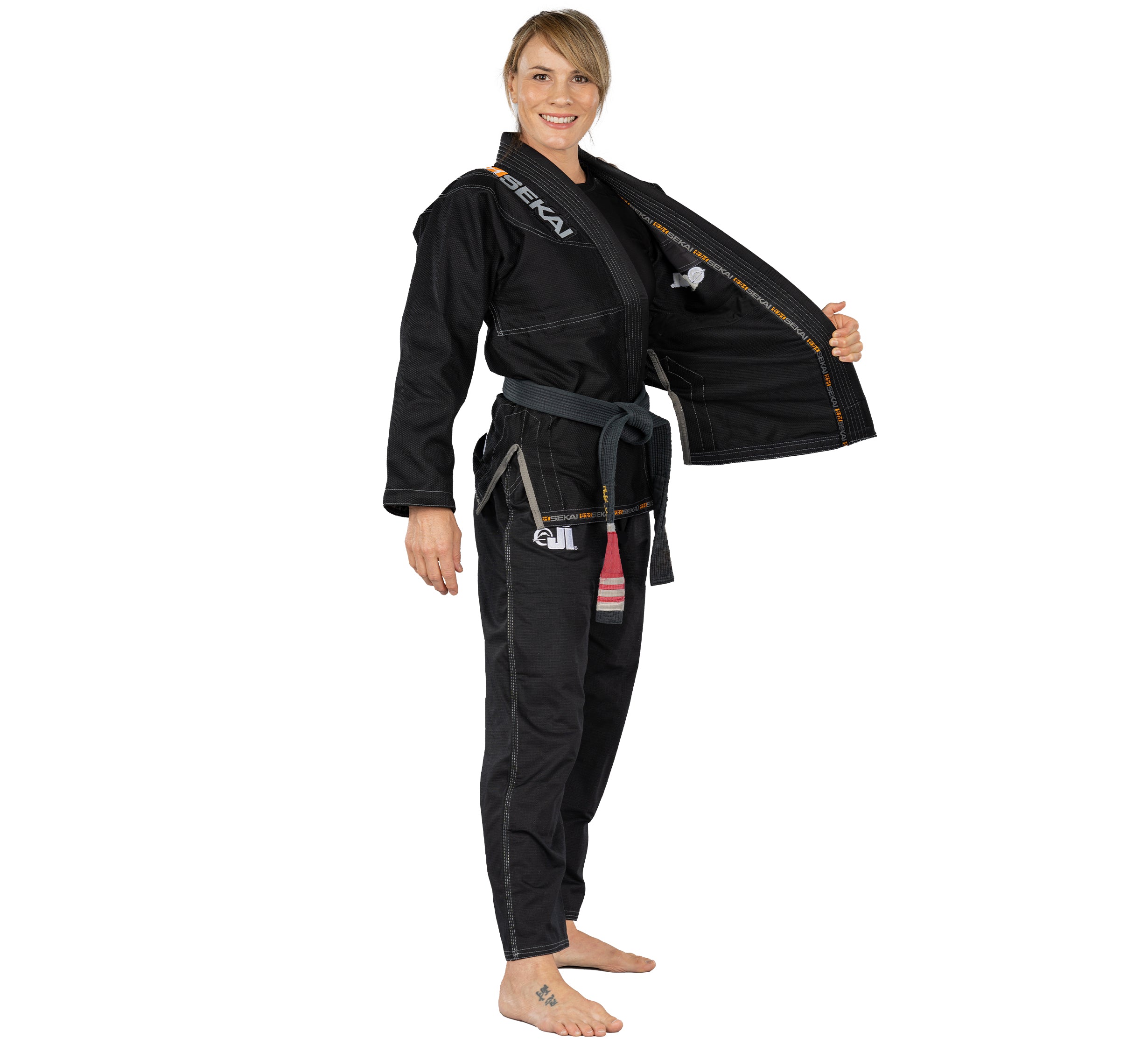 A woman wearing the Sekai 2.0 Womens BJJ Gi Black with a grey belt smiles, barefoot, and holds her gi jacket open to the side against a plain white background.
