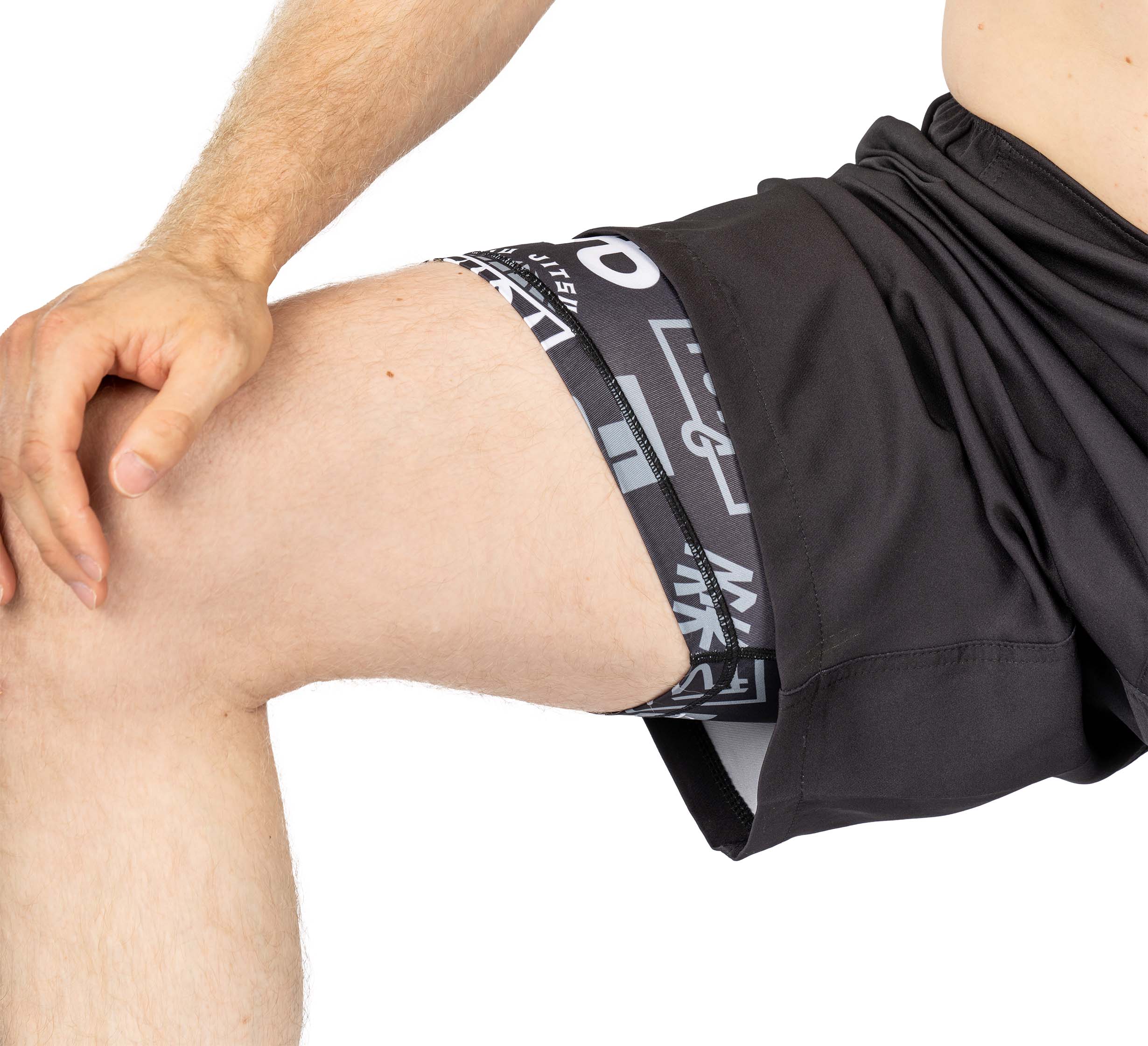 A person wearing Ultimate Grappling Shorts Black lifts one leg to reveal printed compression shorts underneath, with focus on the thigh and shorts as their hand rests on the raised leg.