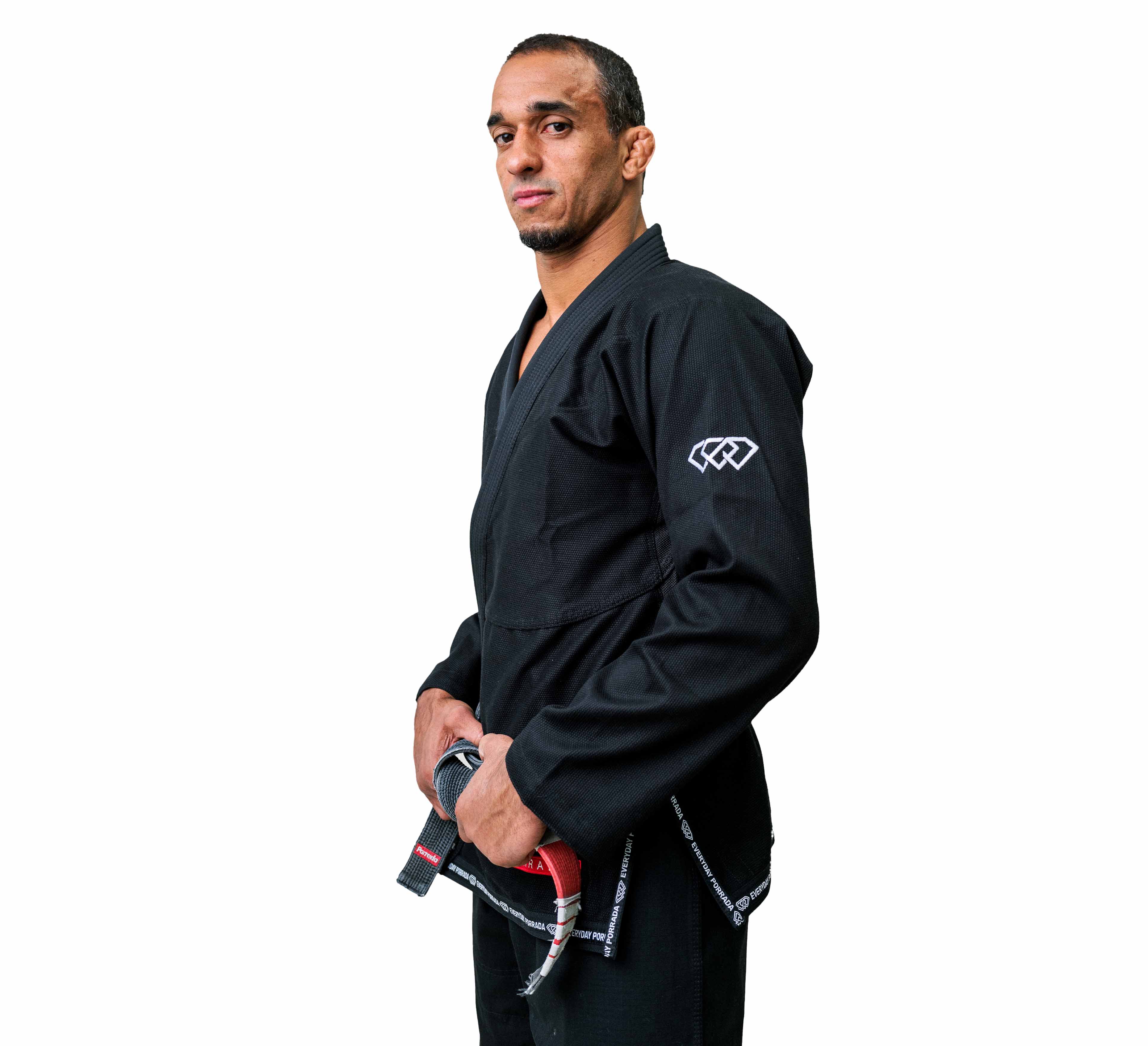 A man wearing the Everyday Porrada Flow-Tech BJJ Gi stands against a white background, confidently holding a black belt with red stripes as he faces forward.