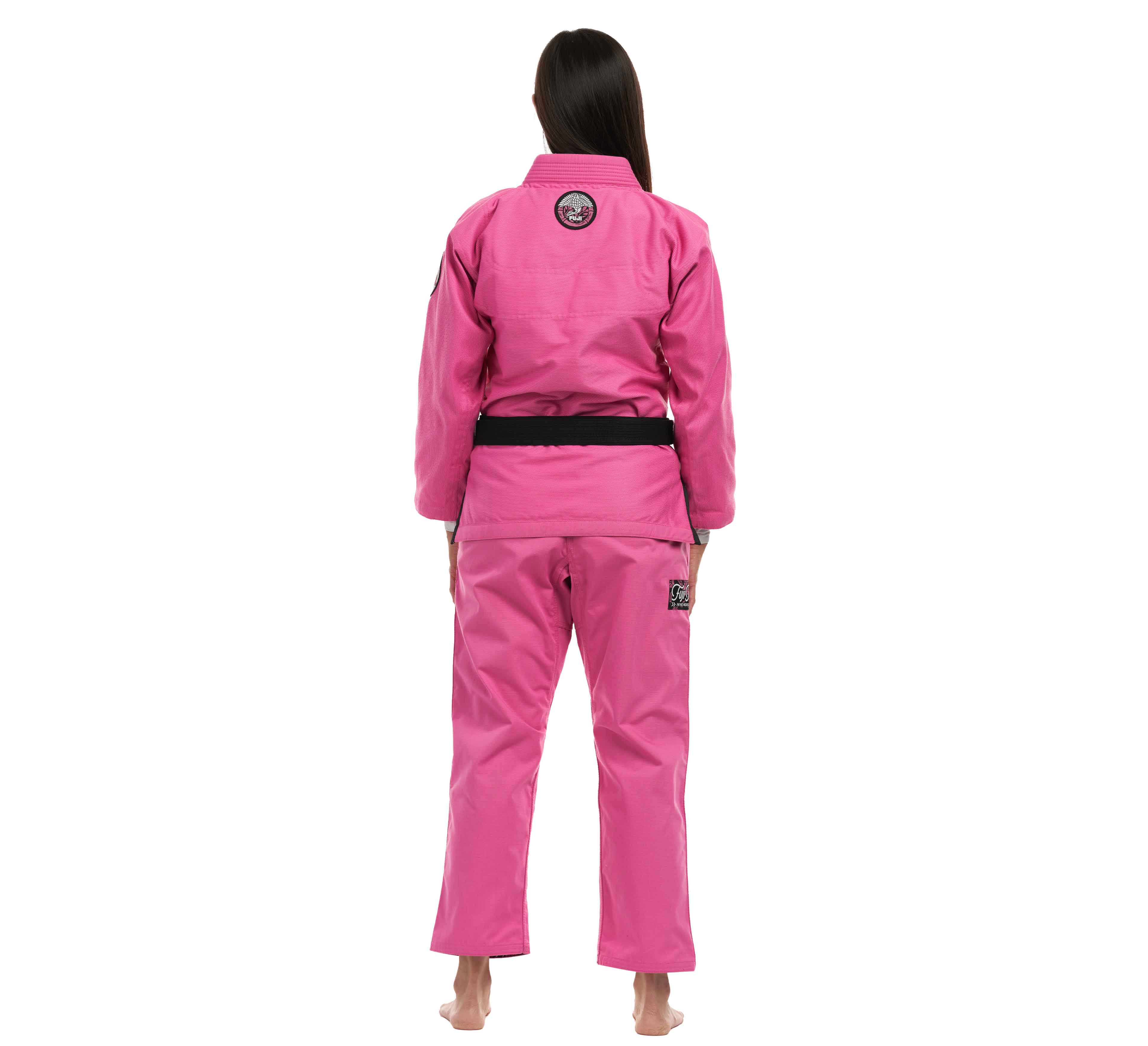A woman with long dark hair wears the Suparaito Womens BJJ Gi Crane Edition in pink with a black belt, standing barefoot and facing away from the camera against a white background.