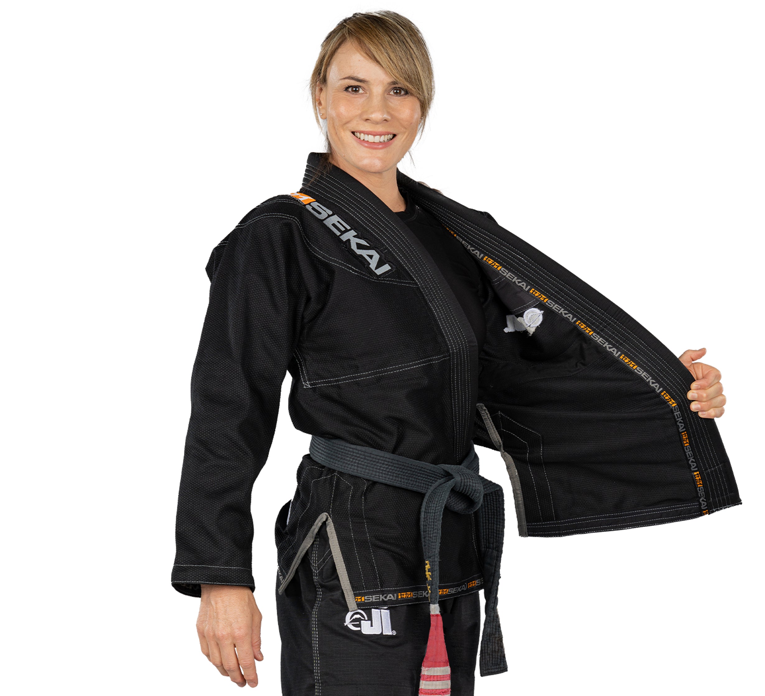 A woman wearing the Sekai 2.0 Womens BJJ Gi Black and a gray belt smiles at the camera, holding her gi open to reveal the inside lining.