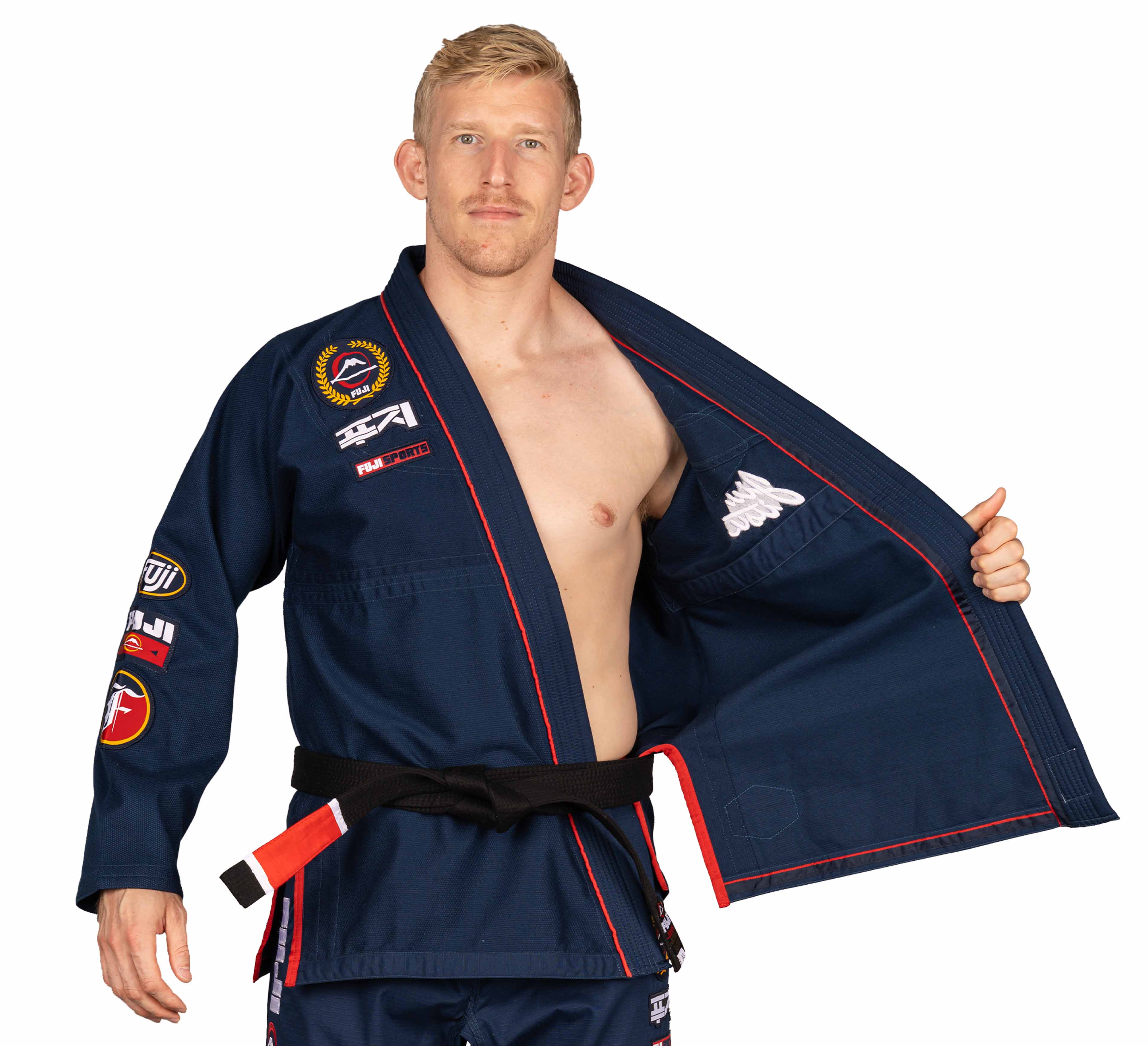 A man with short blond hair, wearing the Suparaito BJJ Gi XTR Edition Navy with patches and a black belt, holds open the left side of his gi to reveal the inner lining.