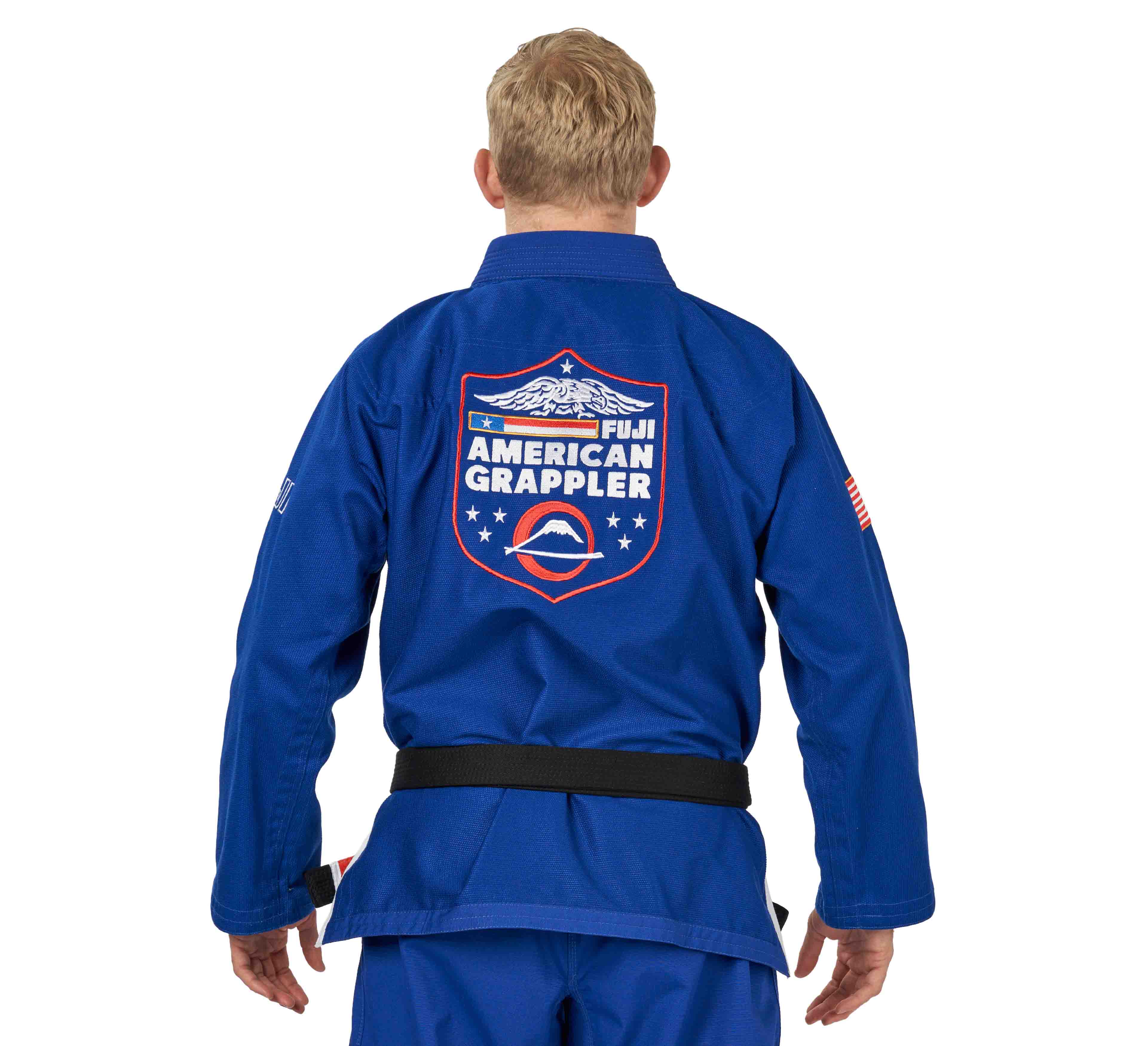 A blond-haired person wears the Suparaito BJJ Gi American Grappler Blue with a black belt. The back displays "Fiji American Grappler" and a logo featuring stars and an eagle.