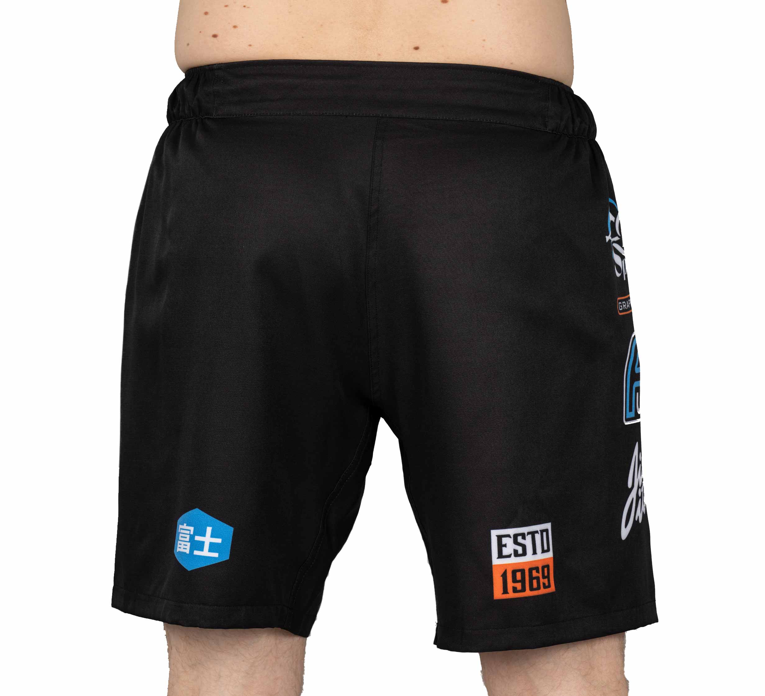 A person stands facing away, showing only their lower back and legs in XTR Extreme Grappling Fight Shorts Black, which feature colorful patches and bold blue, white, and orange graphic elements on the back.