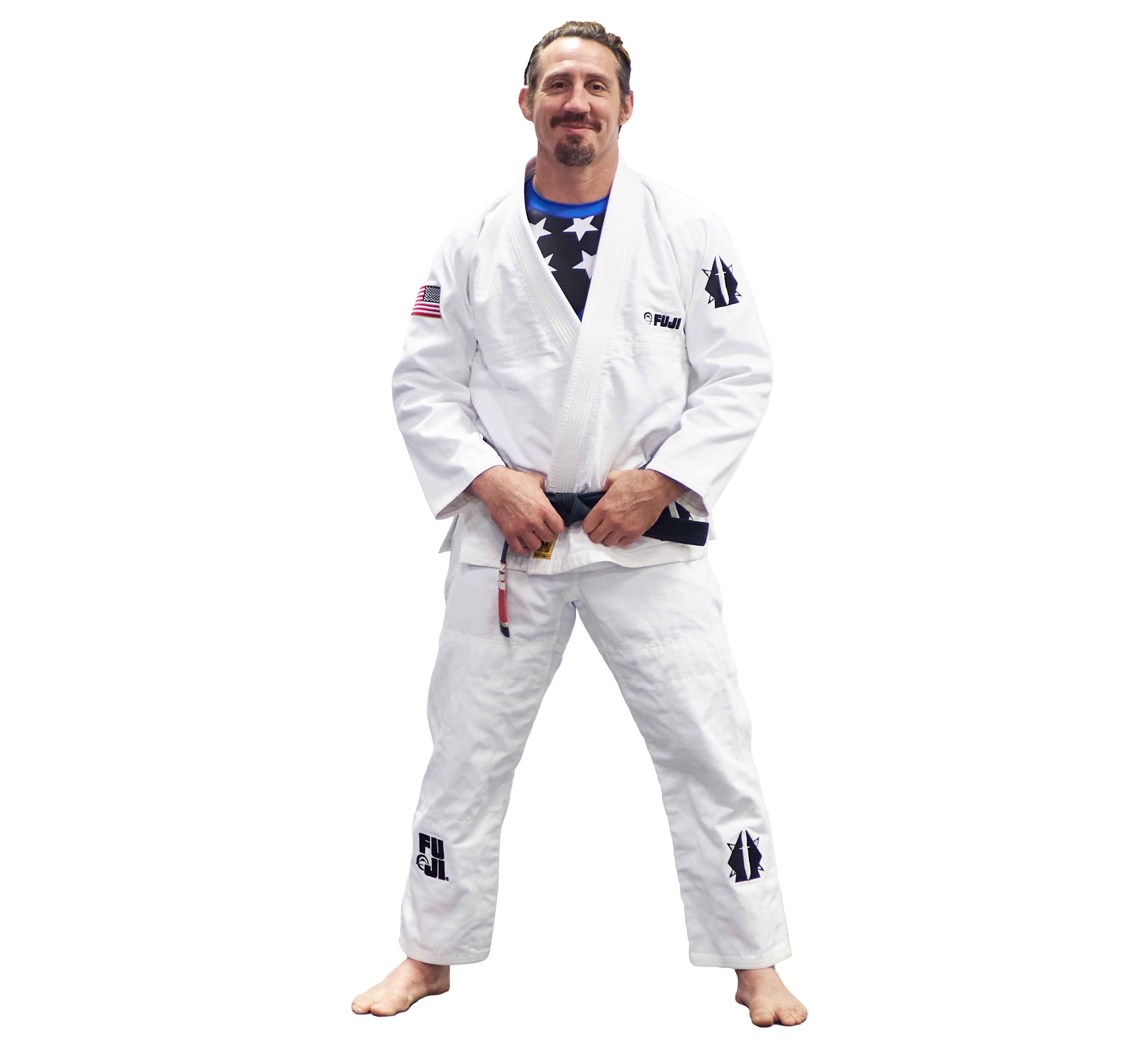 A barefoot, smiling man with a short beard and mustache wears the Lightweight BJJ Gi Sheepdog Response Edition White with patches over a blue rash guard. He holds his belt with both hands.