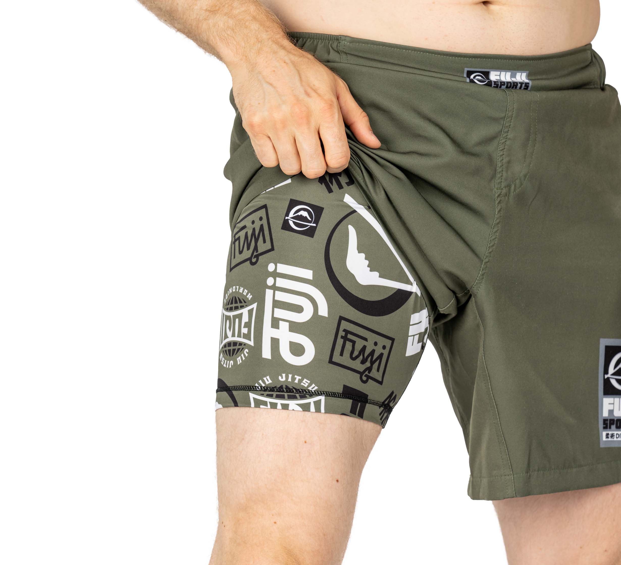 A person lifts the outer layer of their Ultimate Grappling Shorts Military Green to show a compression layer underneath with black and white logos. Only the lower torso and upper legs are visible.