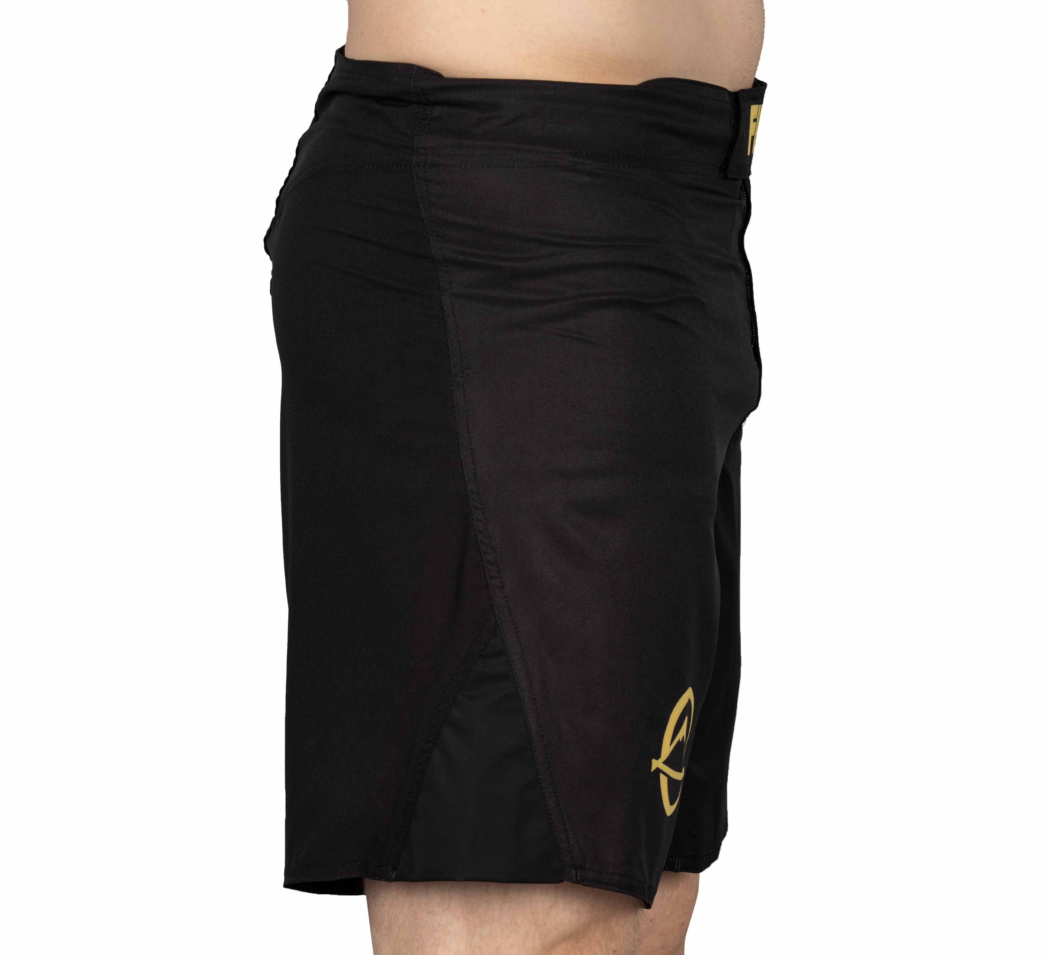 A person wearing Baseline Fight Shorts Black/Yellow, featuring a small yellow logo near the bottom hem, is shown from the side against a white background.