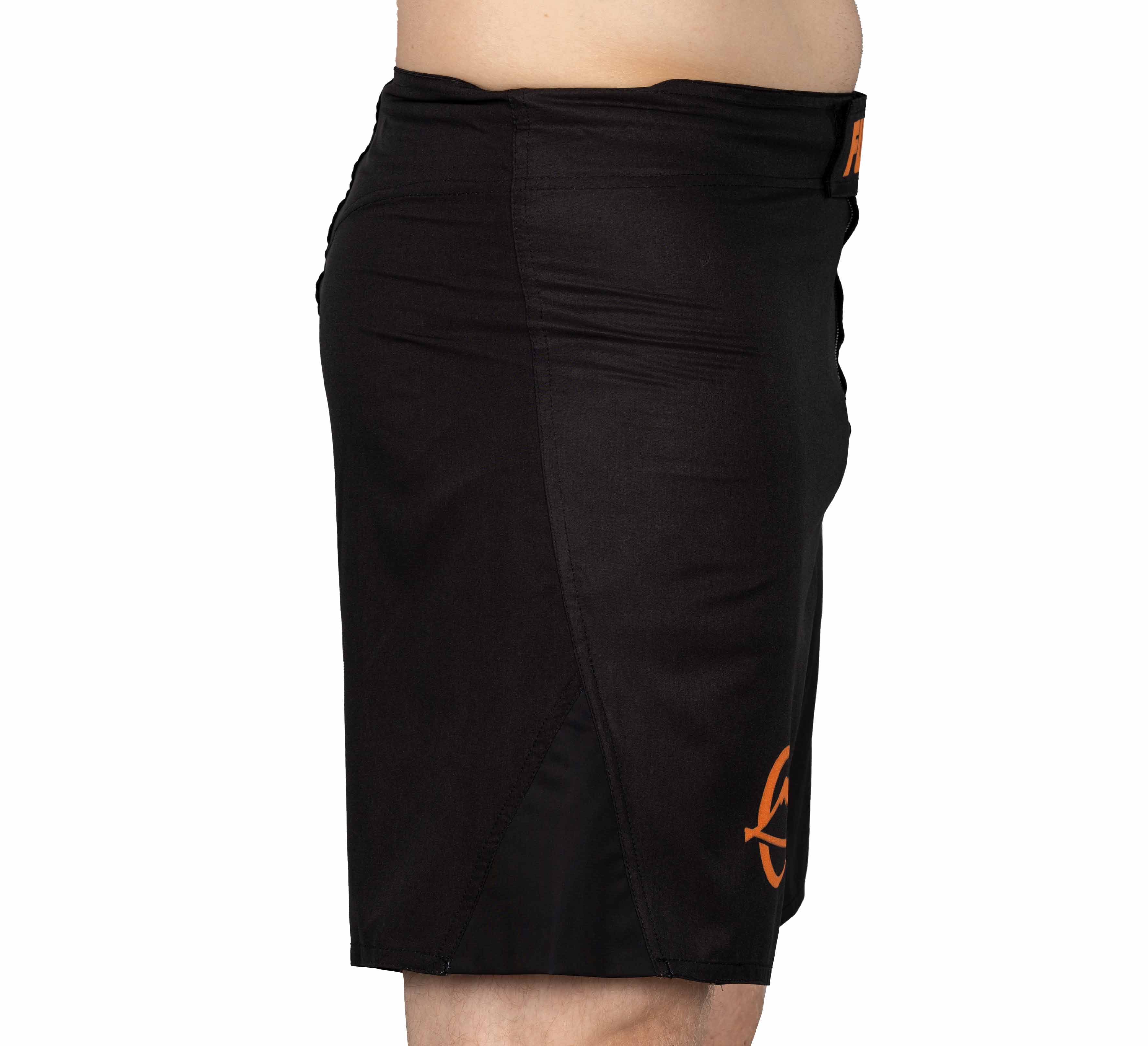 A person in Baseline Fight Shorts Black/Orange featuring an orange logo on the lower leg, shown from the side against a white background.