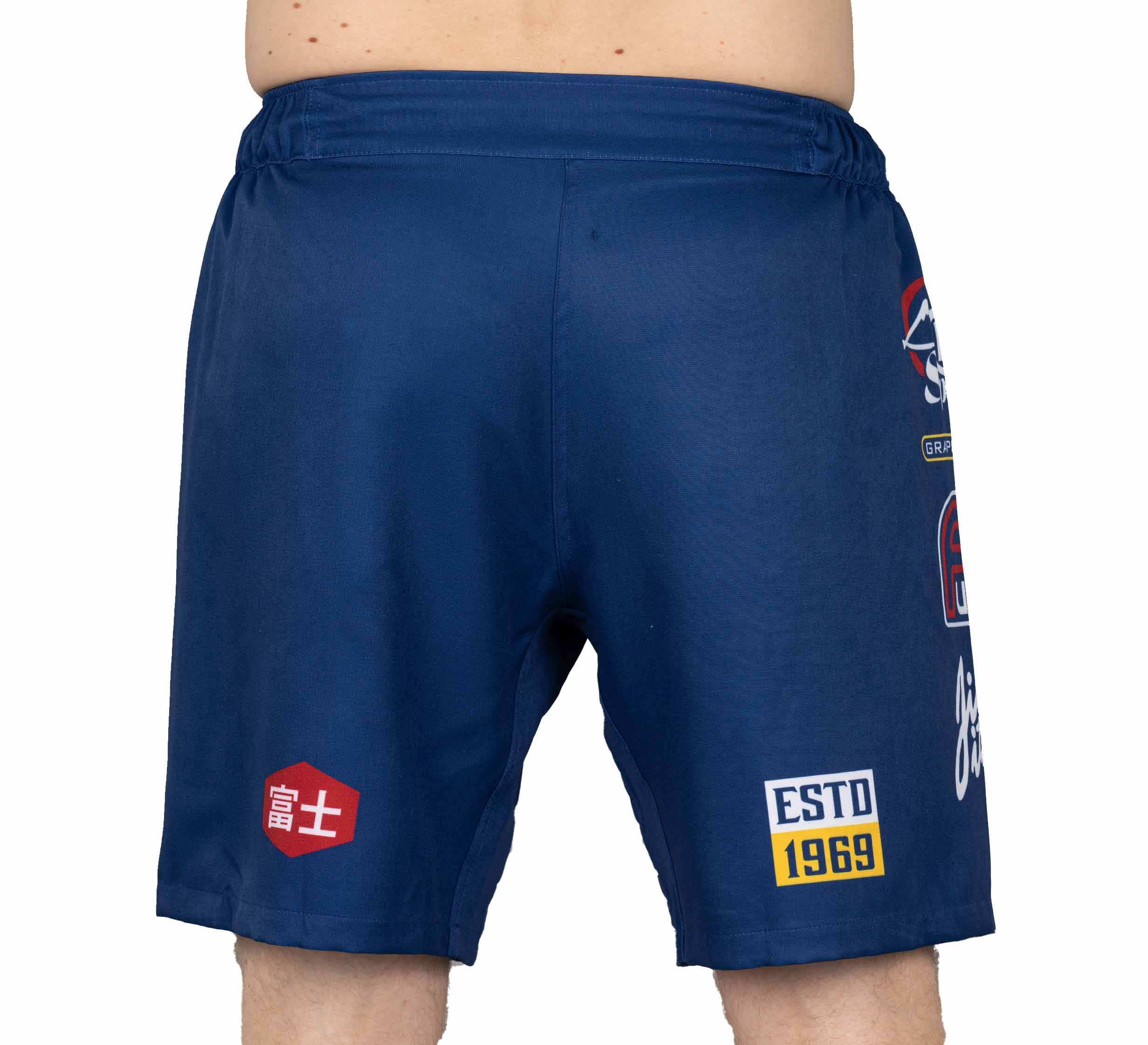Someone shown from the back wears XTR Extreme Grappling Fight Shorts Navy, featuring various patches and graphics, including a red hexagon with Japanese characters, a yellow ESTD 1969 patch, and other multicolored logos.