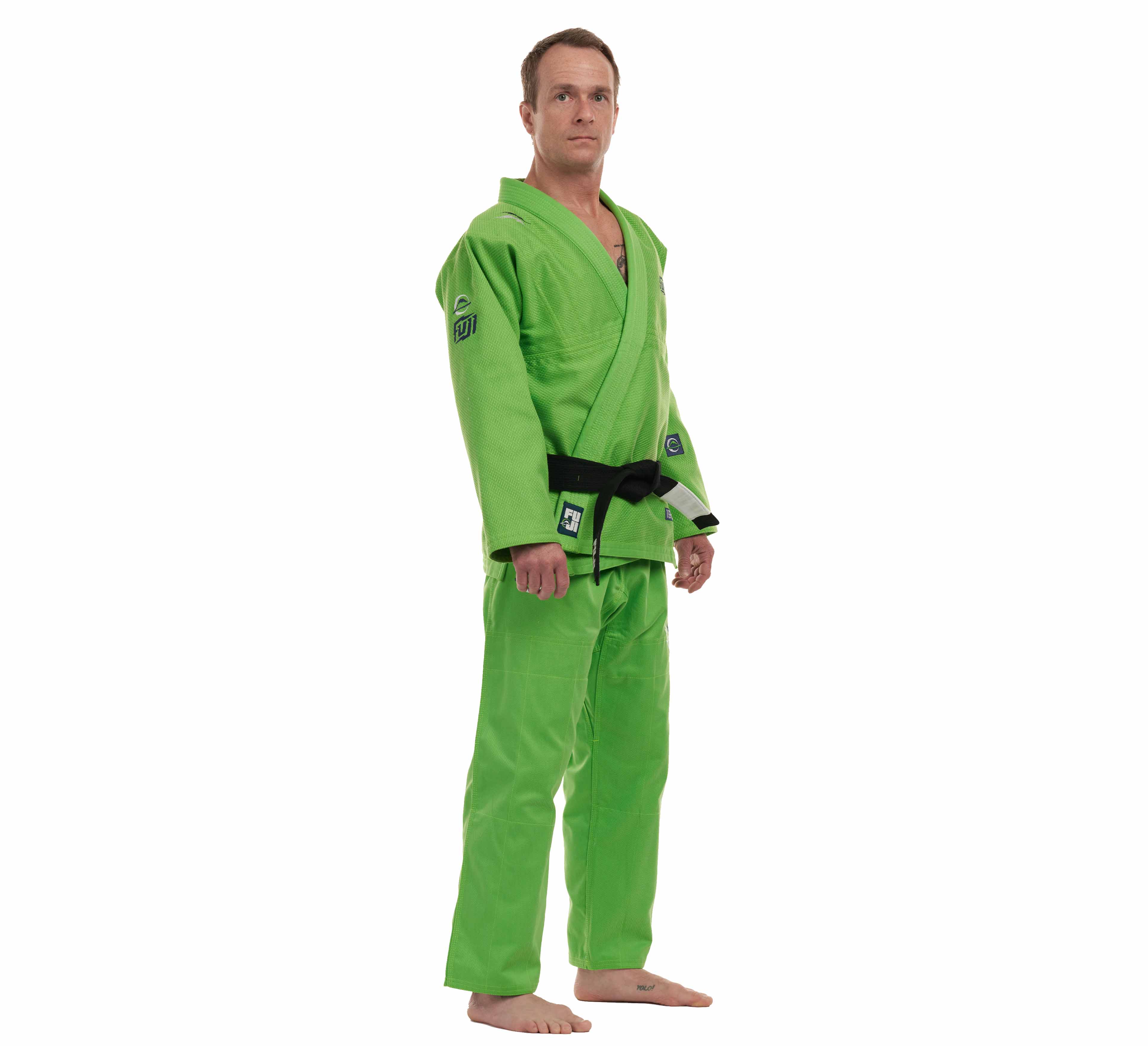 A man stands barefoot, wearing the All Around BJJ Gi Electric Green Edition with a black belt. He faces forward with a neutral expression against a plain white background.