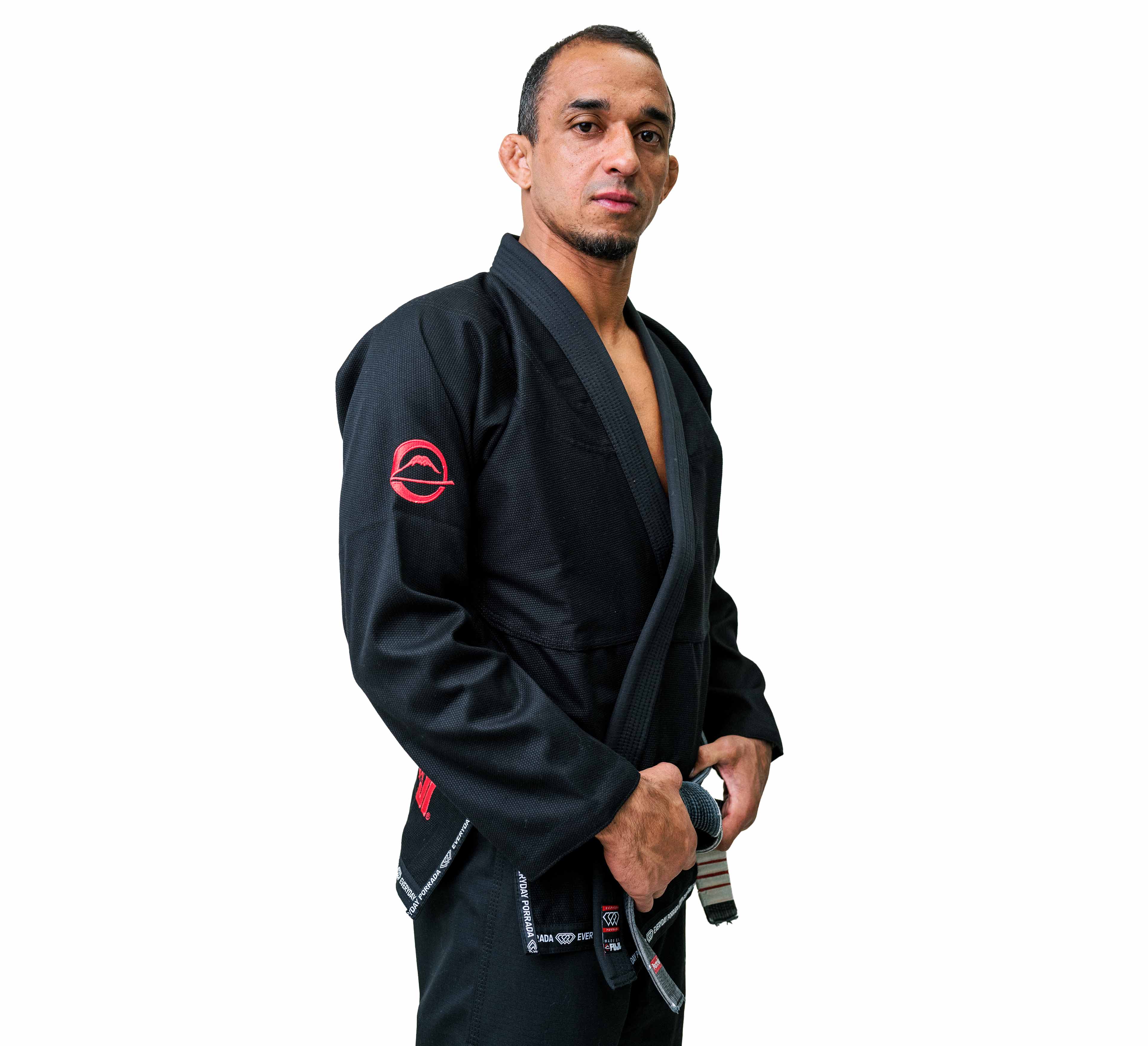 A man stands against a white background, confidently holding his black belt while wearing the Everyday Porrada Flow-Tech BJJ Gi, which features distinctive red patches.