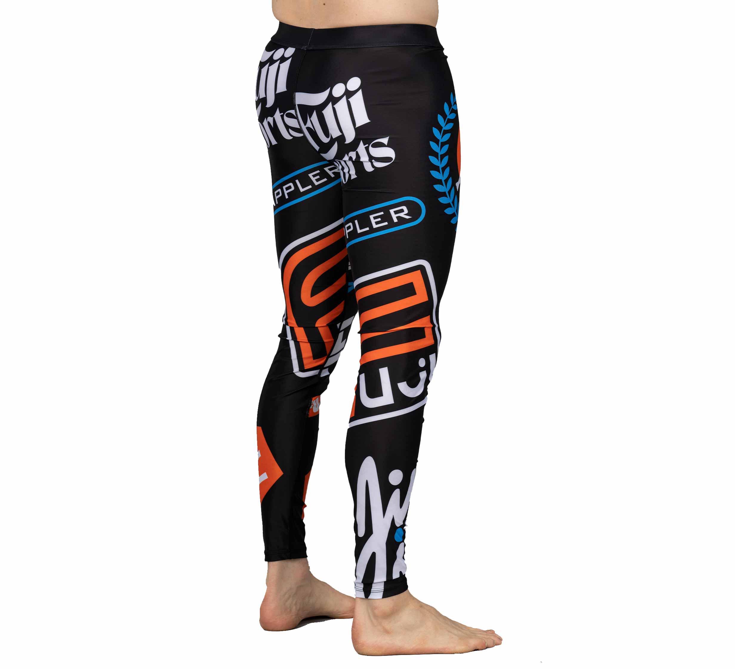 Model wears XTR Extreme V1 Grappling Spats Black—fitted black leggings with bold logos and text in white, orange, and blue. Barefoot, the model stands against a white background.