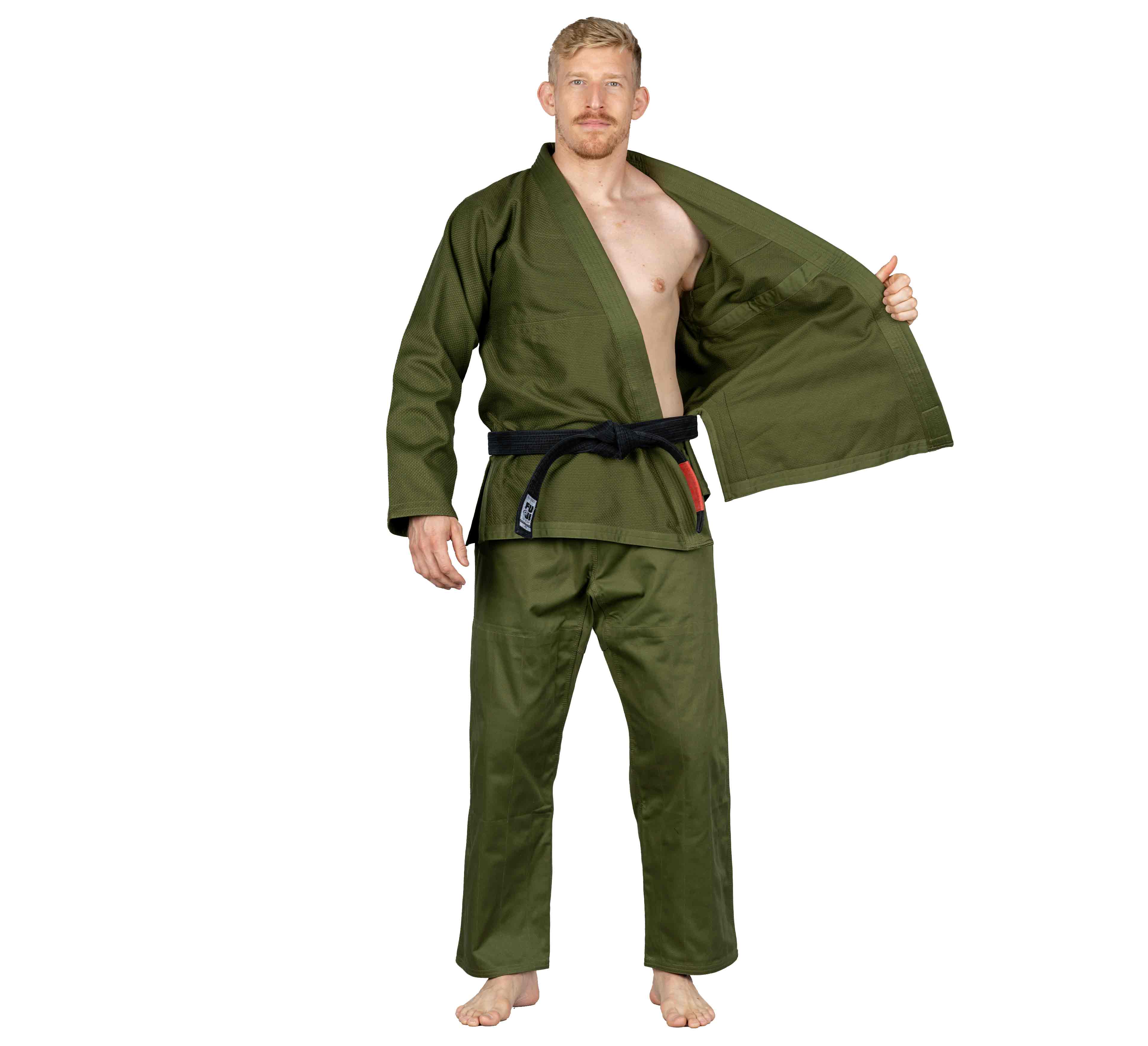 A man with short blond hair and a beard wears the All Around BJJ Gi in Military Green with a black belt, standing barefoot against a white background and holding one side of his open jacket.