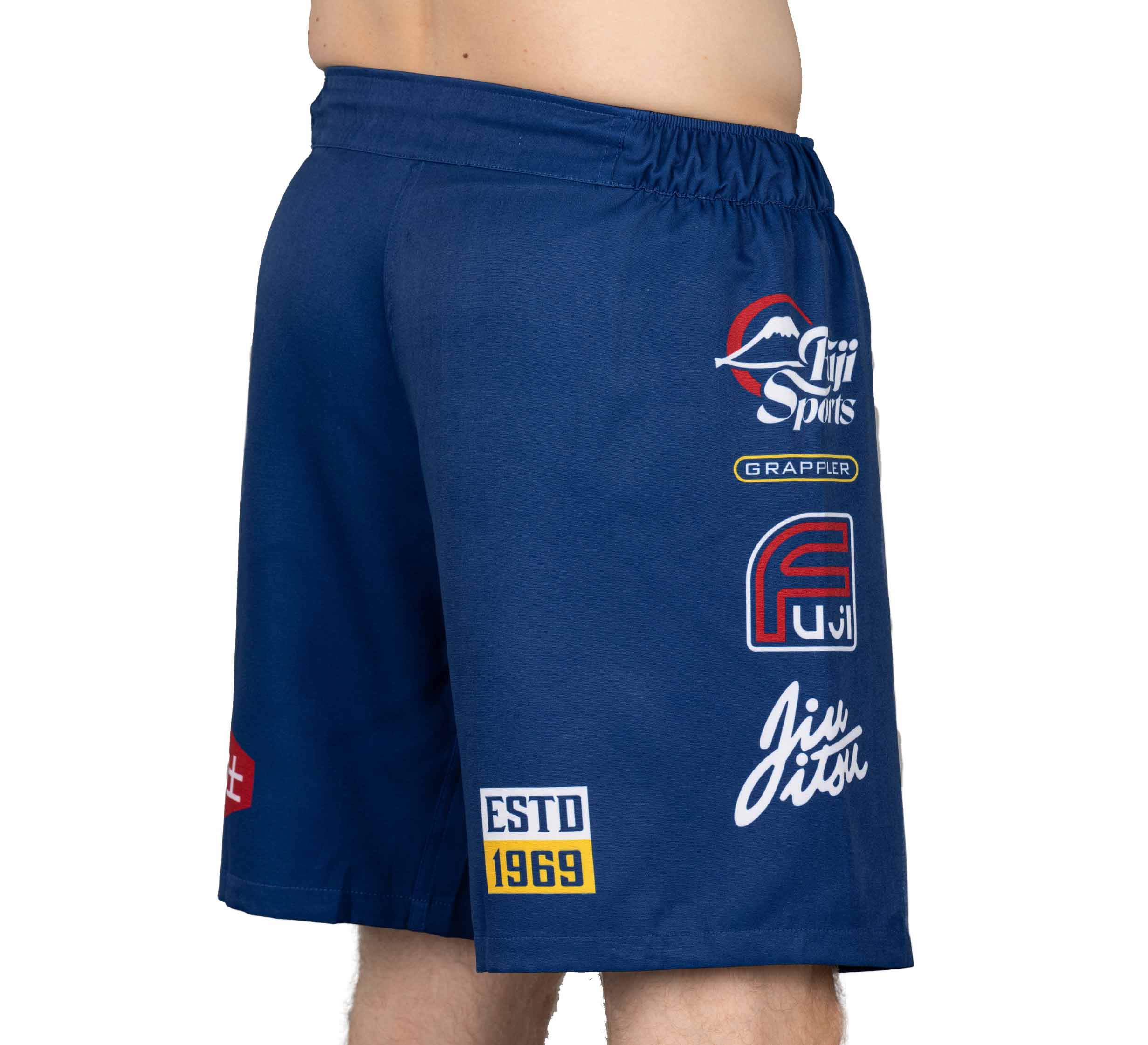 A person wears XTR Extreme Grappling Fight Shorts Navy with multiple logos and patches—including Fuji Sports, Grappler, Fuji, ESTD 1969, and Jiu Jitsu—on the left side; only the lower torso and upper legs are visible.