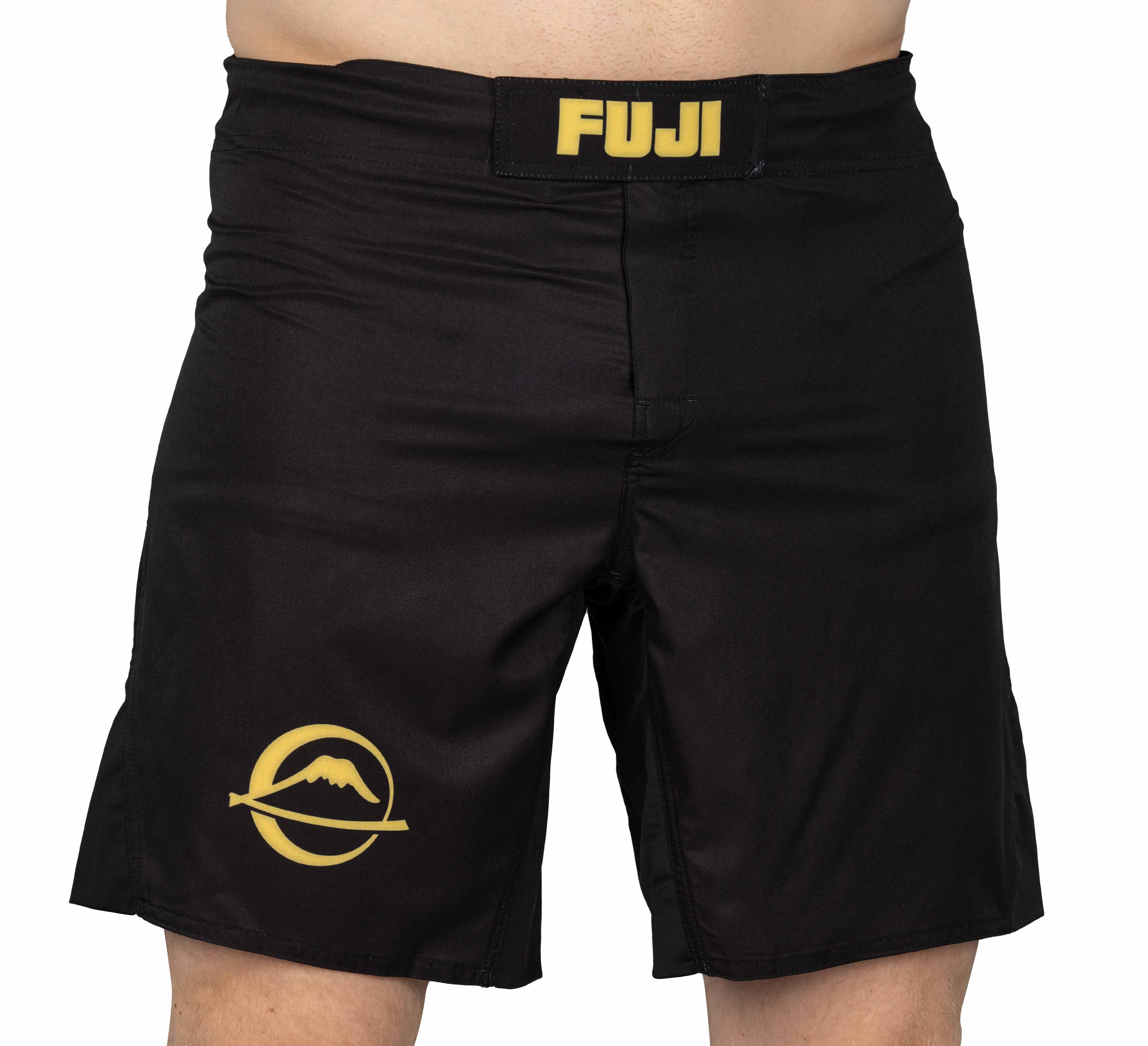 A person wearing Baseline Fight Shorts Black/Yellow featuring a yellow FUJI logo on the waistband and a yellow mountain logo on the left leg.