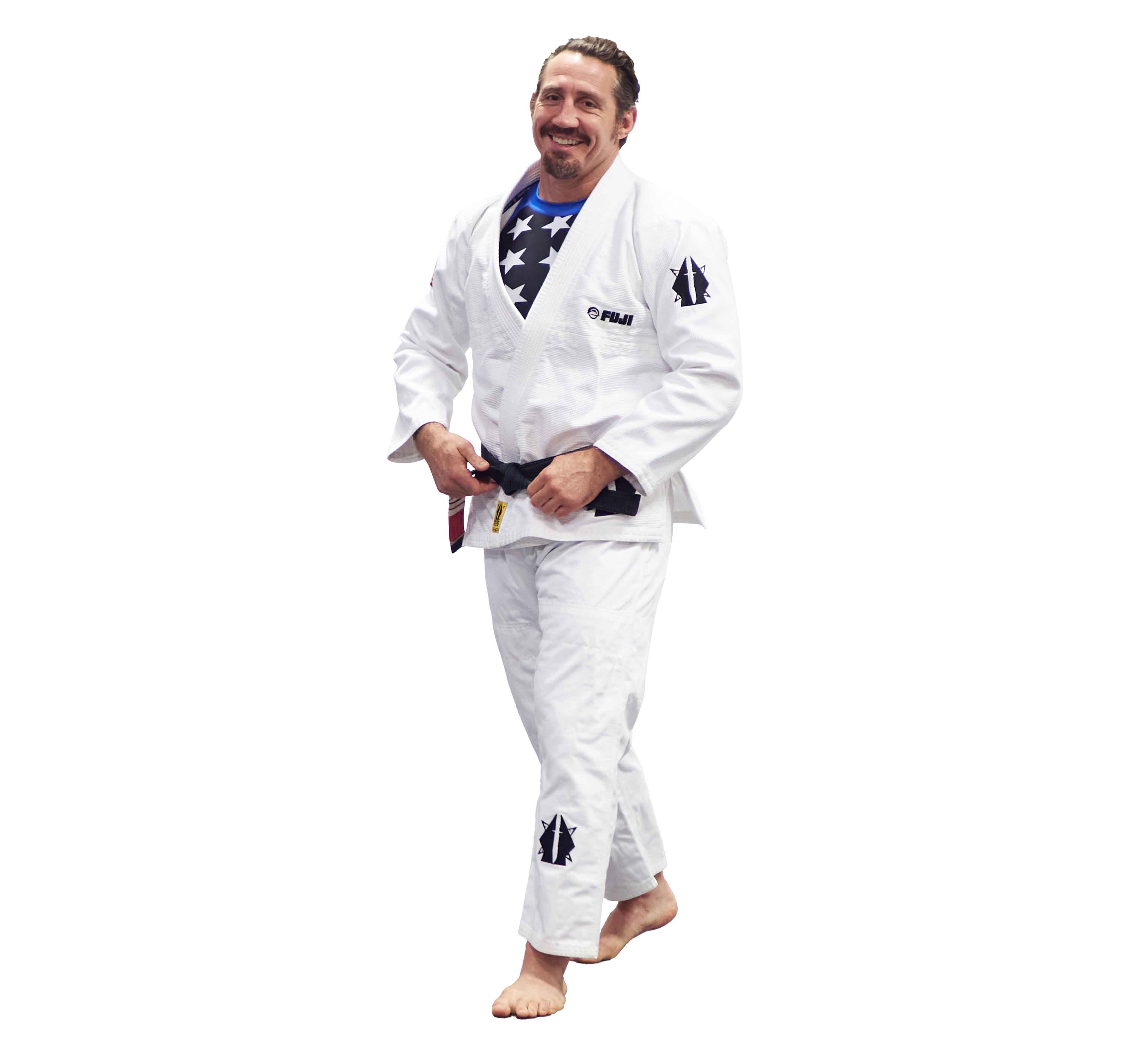 A man, barefoot and smiling, stands against a white background in the Lightweight BJJ Gi Sheepdog Response Edition White with black patches, a black belt, and a blue shirt with white stars underneath.
