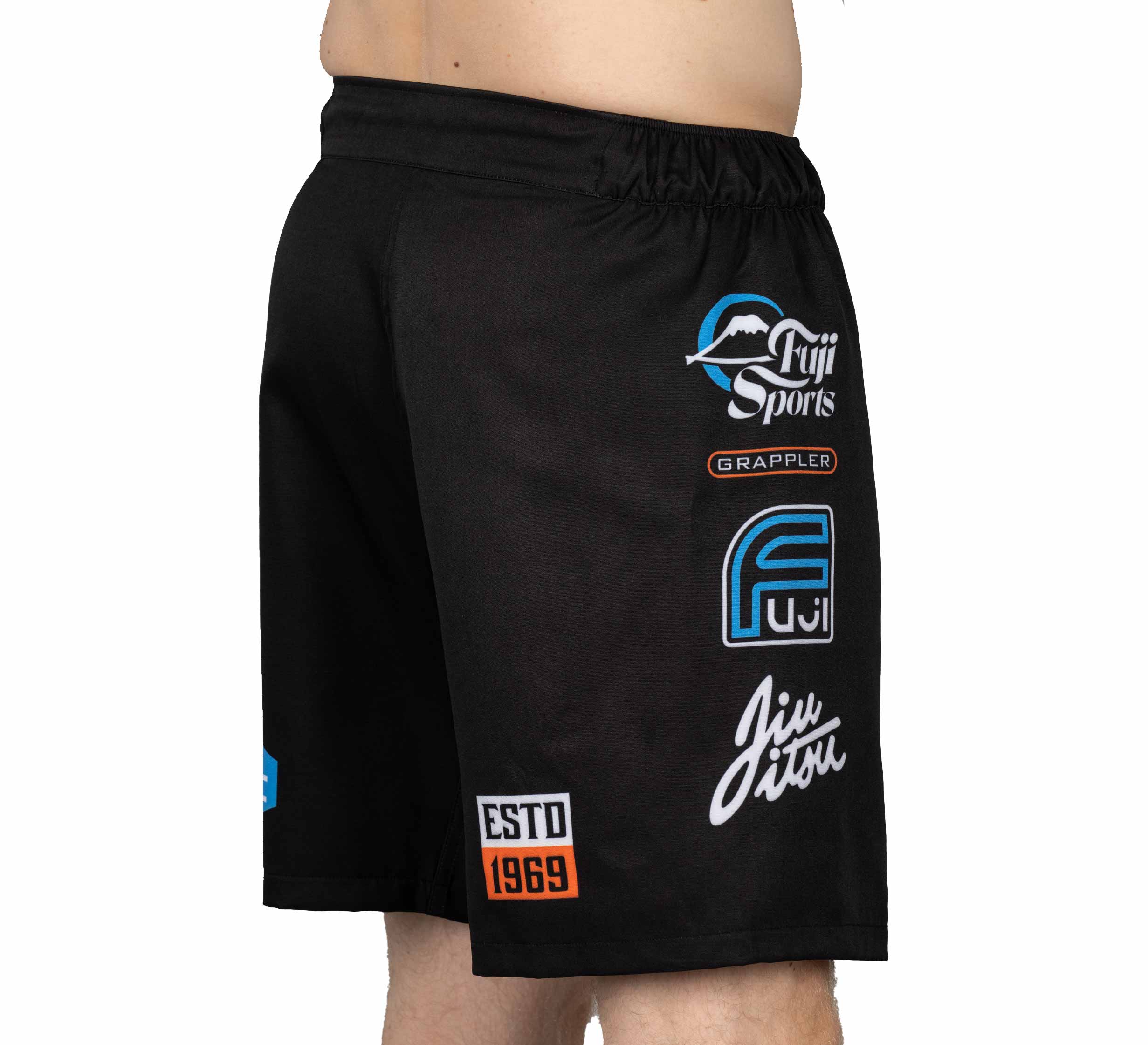 A person wearing XTR Extreme Grappling Fight Shorts Black, featuring Fuji Sports, Grappler, Jiu Jitsu, ESTD 1969 logos and a blue F symbol; shown from waist to mid-thigh in side view.