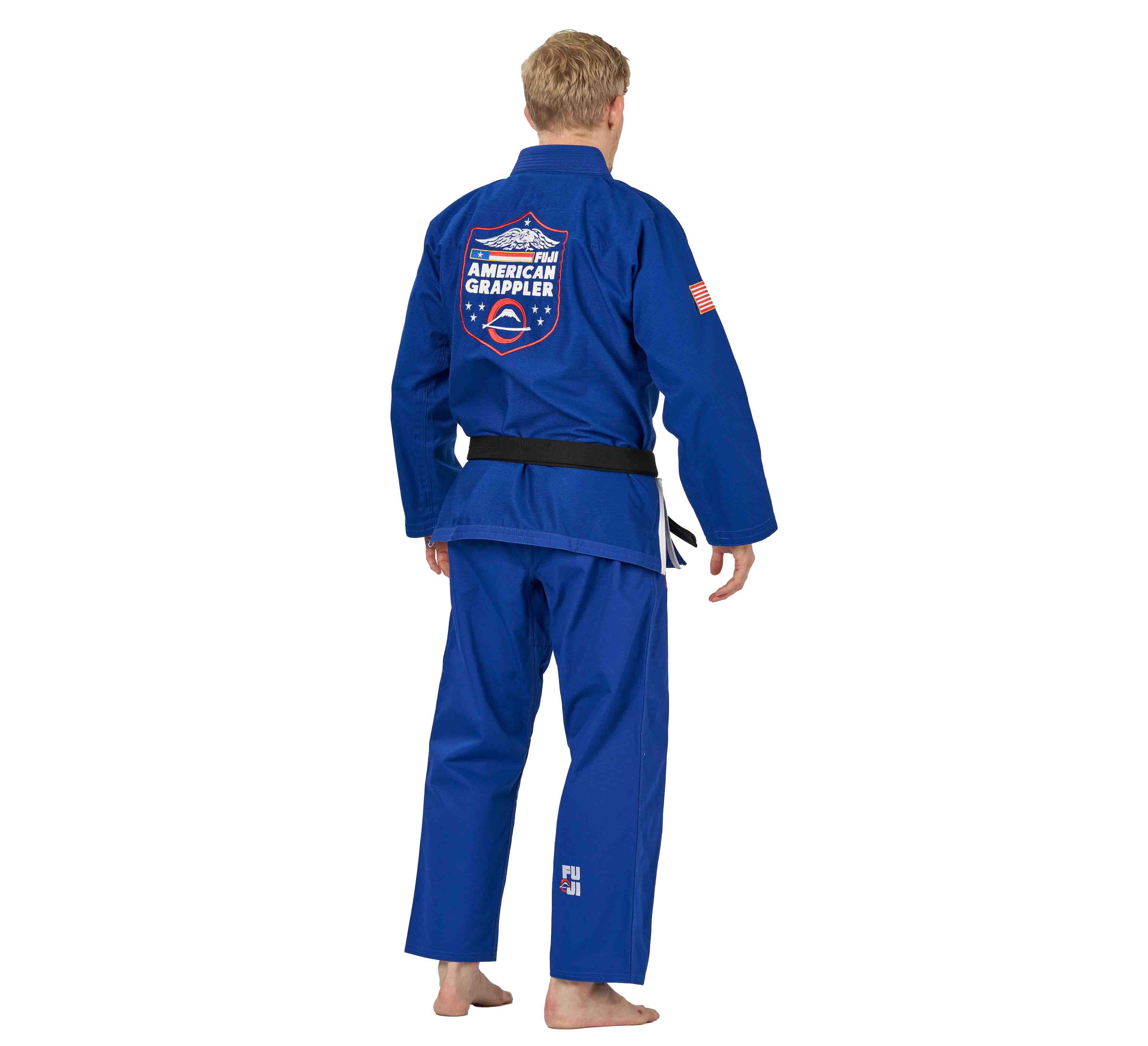 A barefoot person with short blond hair faces away, wearing the Suparaito BJJ Gi American Grappler Blue with a black belt. A large American Grappler patch is on the back and a small U.S. flag appears on the upper sleeve of the gi.