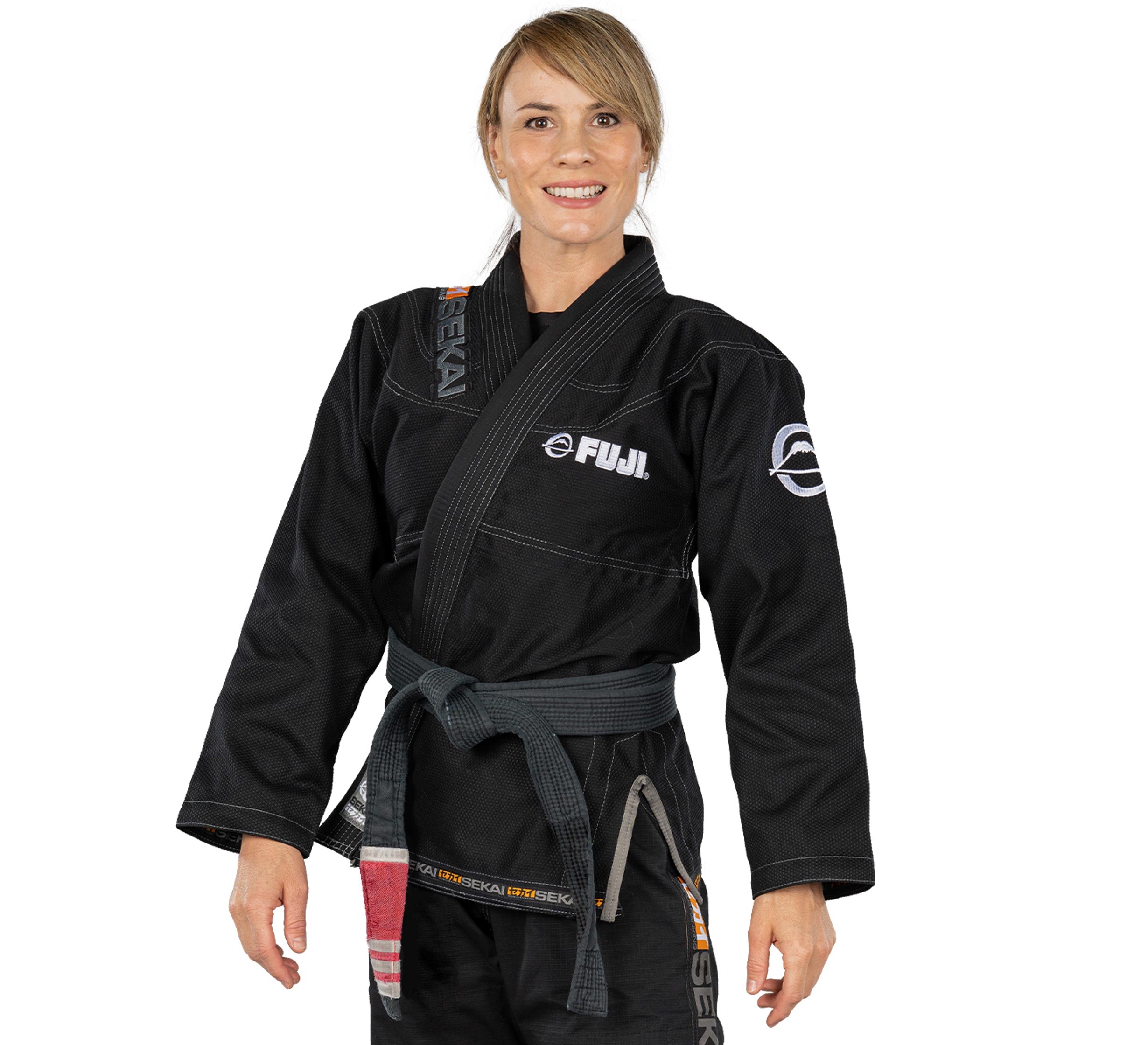 A woman stands smiling, wearing the Sekai 2.0 Womens BJJ Gi Black with FUJI logos and a grey belt tied at her waist.