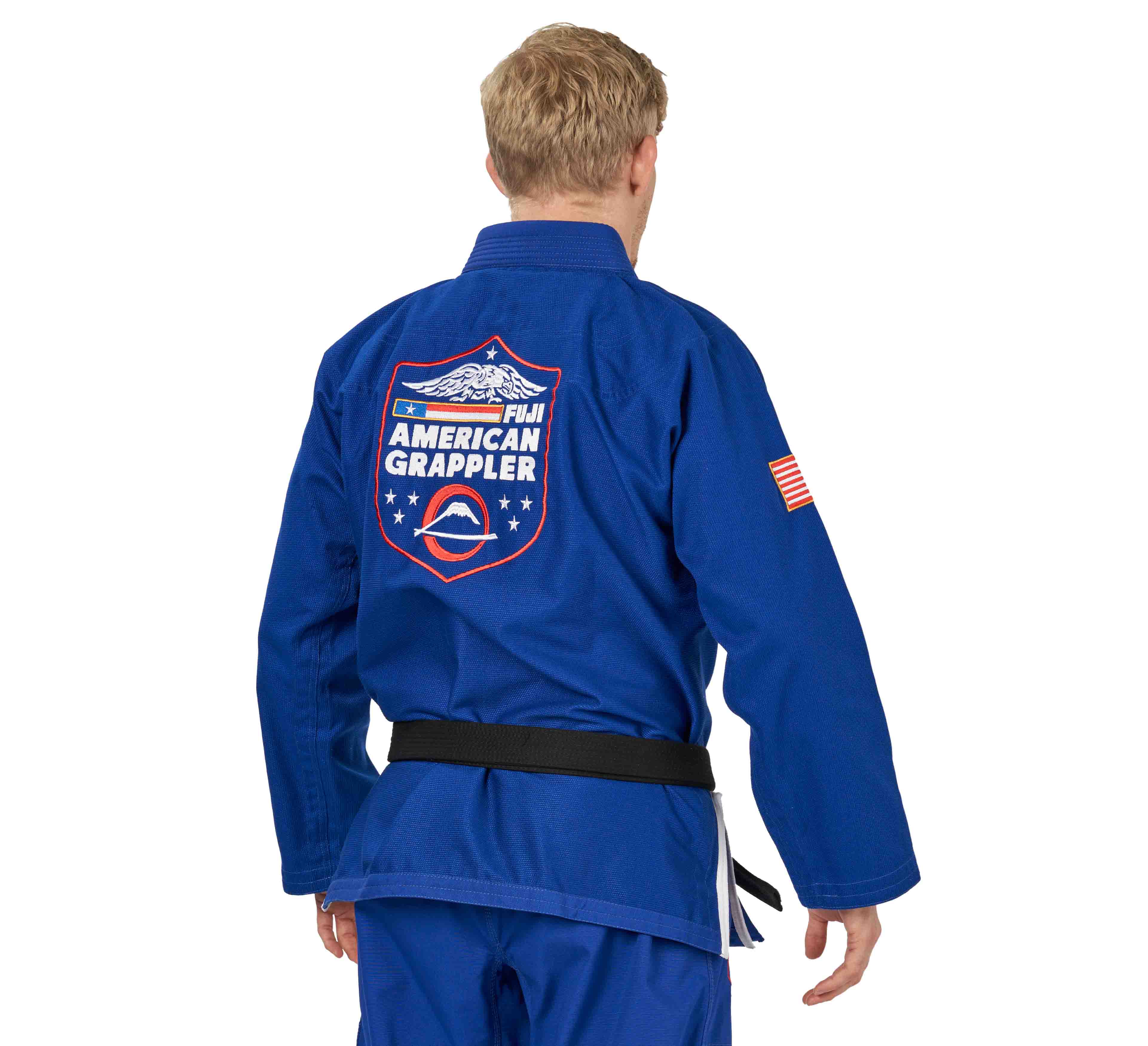 A person is shown from behind wearing the Suparaito BJJ Gi American Grappler Blue, featuring a mountain logo and "American Grappler" on the back, plus an American flag patch on the sleeve.