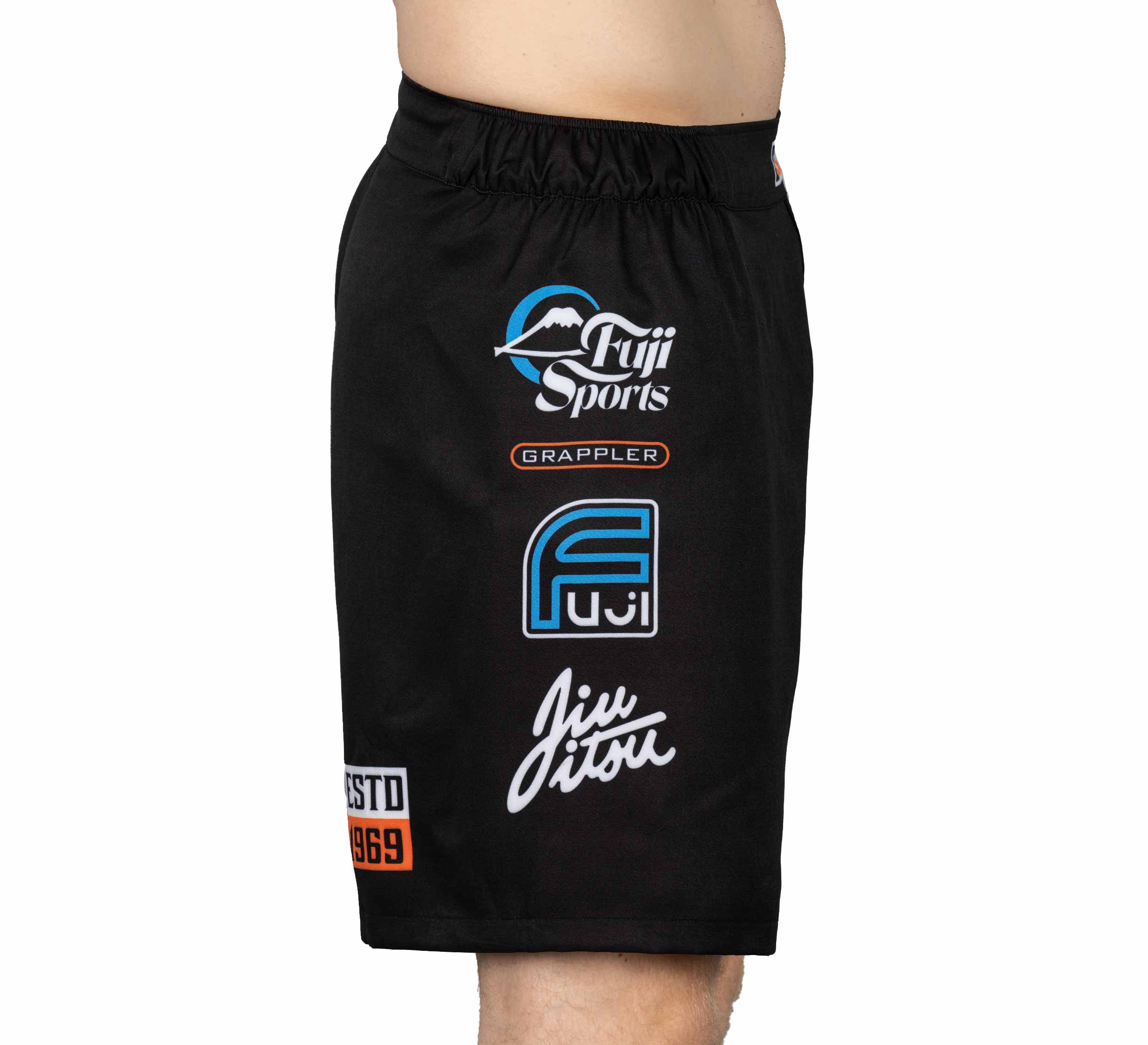 A person in XTR Extreme Grappling Fight Shorts Black, featuring white and blue Fuji Sports and Grappler logos, standing in side profile.