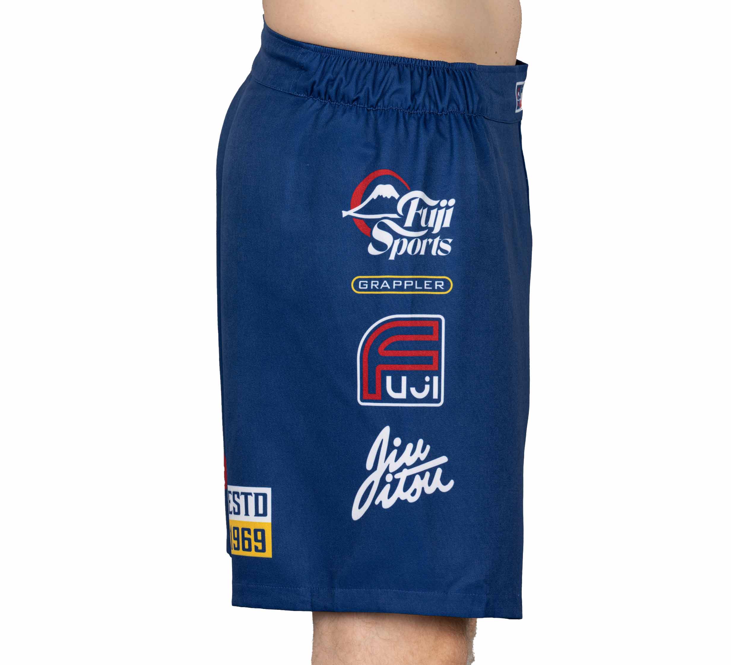 A person wearing XTR Extreme Grappling Fight Shorts Navy featuring white, red, and yellow logos and text such as Fuji Sports, Grappler, Fuji, Jiu Jitsu, and ESTD 1969 on the side.