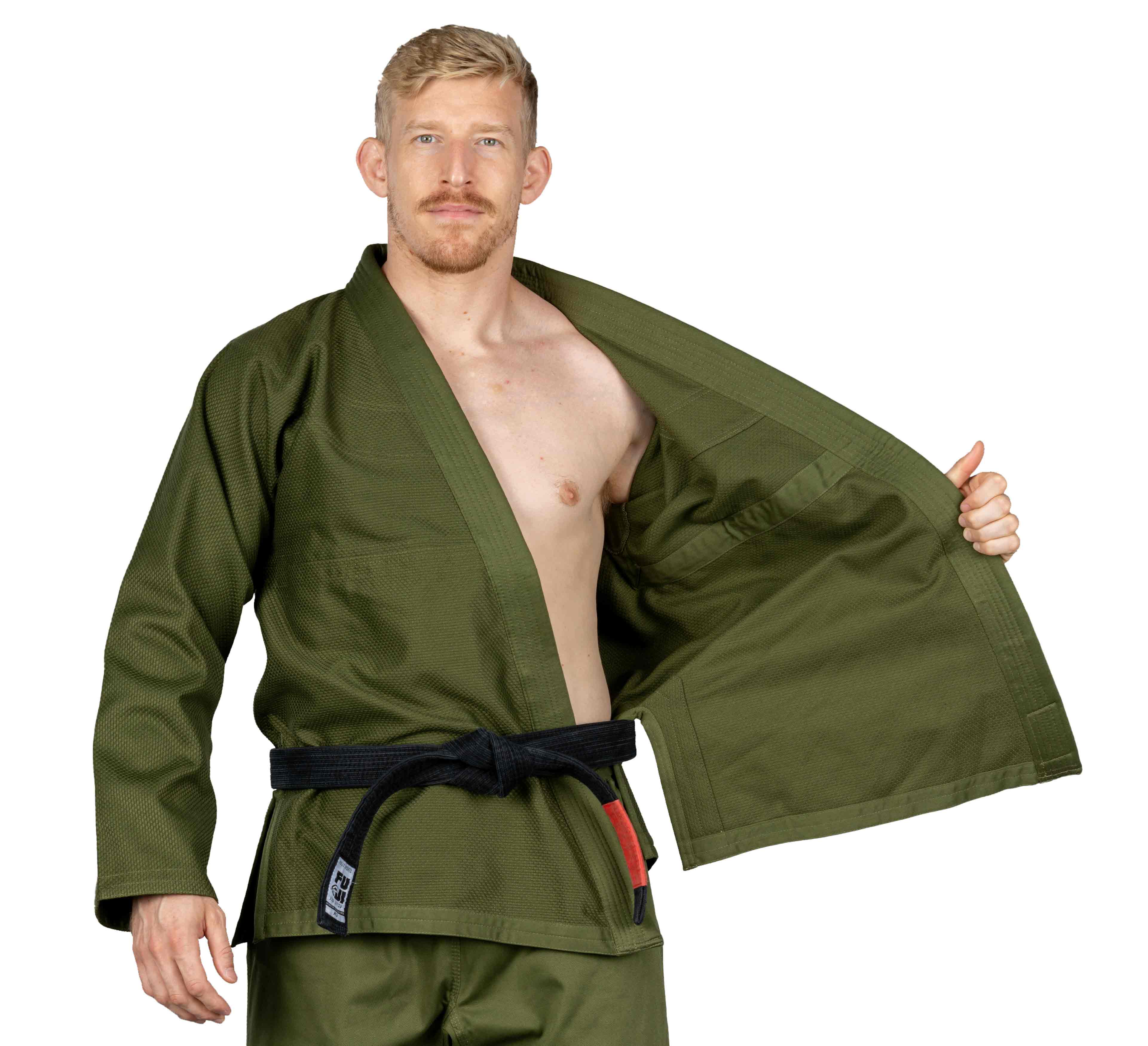 A man wearing the All Around BJJ Gi Military Green with a black belt stands facing forward, opening one side of his jacket to reveal the interior. He has short blond hair, a mustache, and a trimmed beard. The background is plain white.