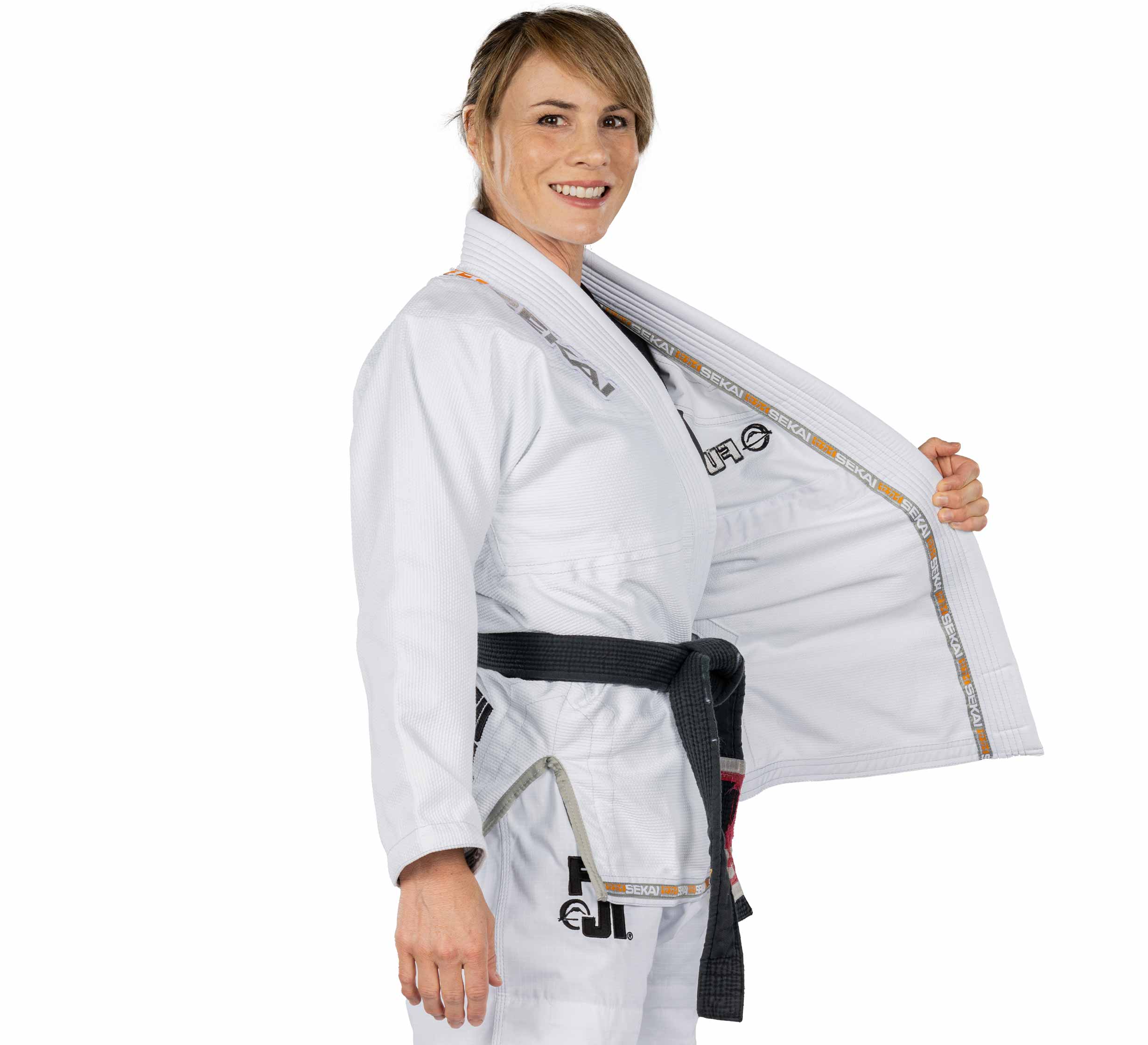 A woman wearing the Sekai 2.0 Womens BJJ Gi White with a black belt smiles and holds open one side of her jacket to show the inside, standing against a white background.