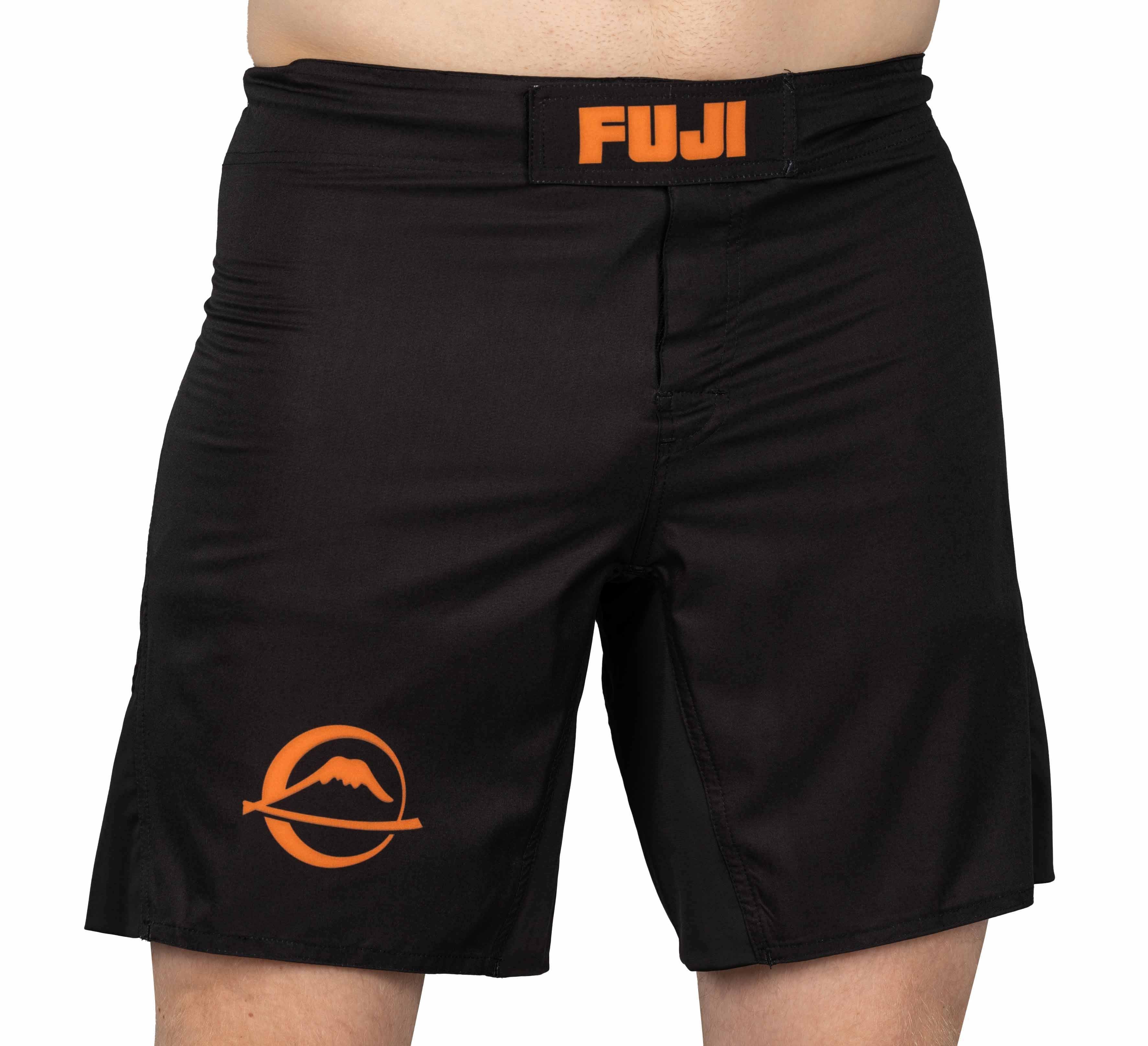 A person wearing Baseline Fight Shorts Black/Orange featuring an orange FUJI logo on the waistband and an orange mountain logo on the left leg.