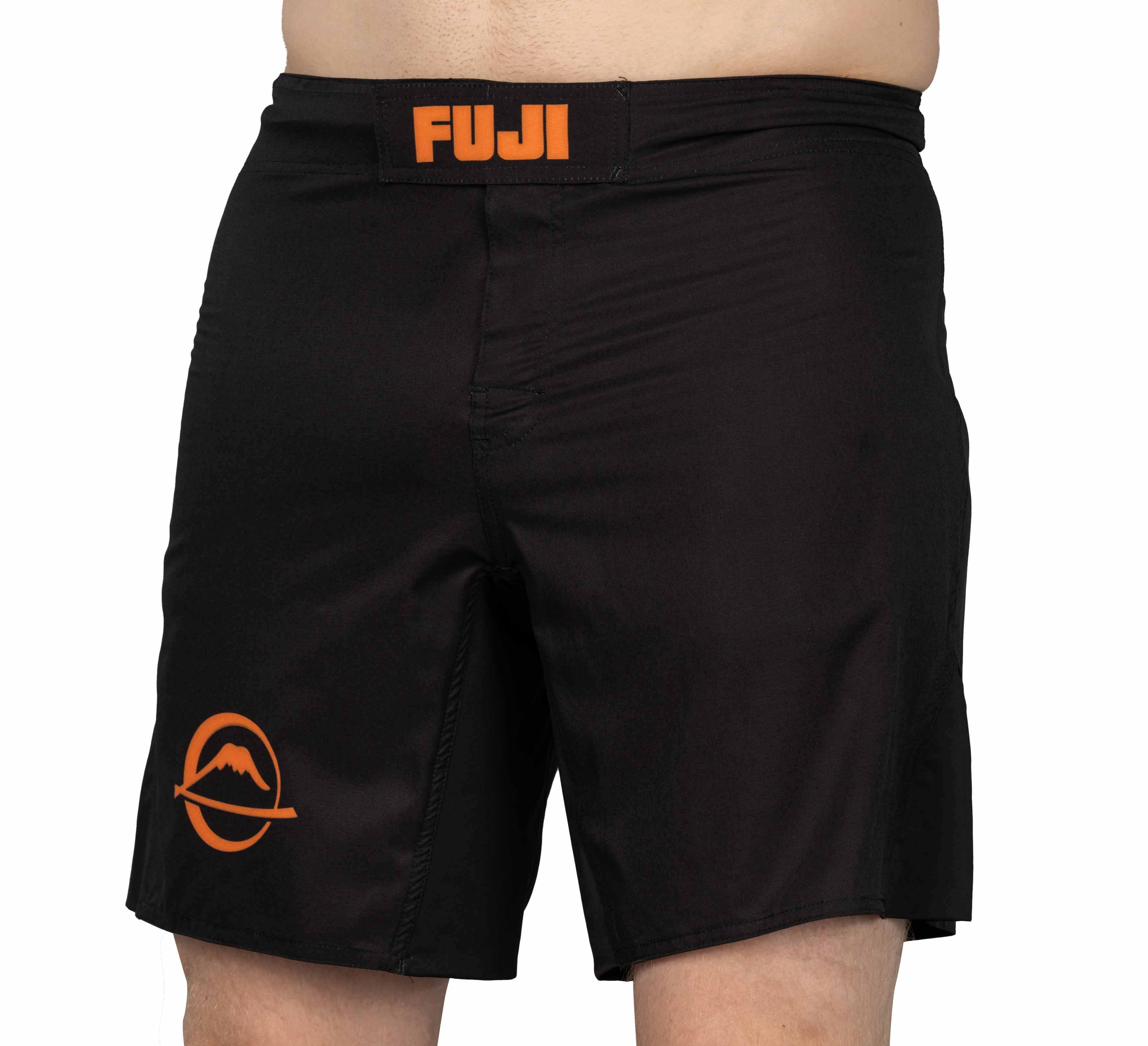 A person is wearing Baseline Fight Shorts Black/Orange, featuring "FUJI" in orange on the waistband and an orange mountain logo on the left thigh. Only the shorts are visible; their upper body and lower legs are not shown.