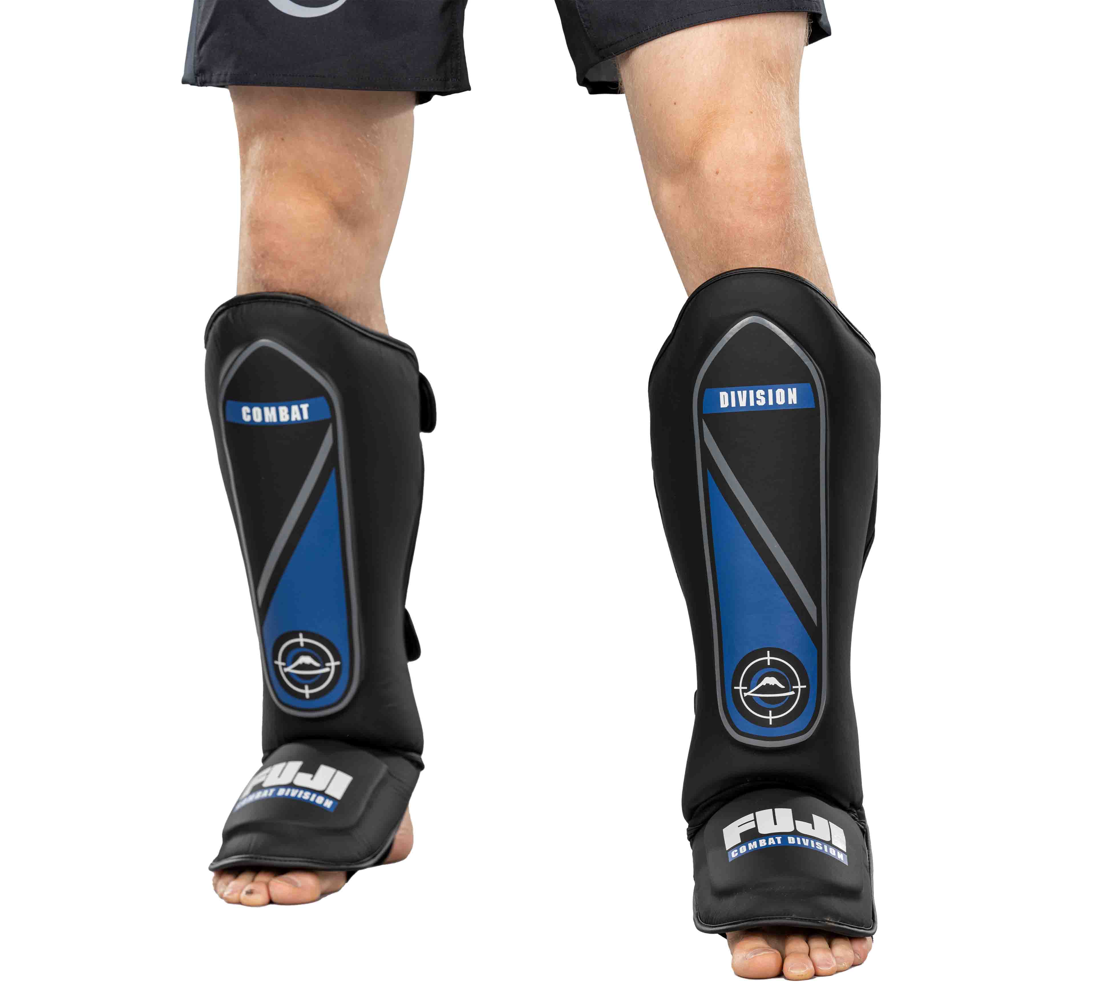 A person wearing black Precision Striking Shin Guards with blue accents, the words COMBAT and DIVISION printed on them, and the FUJI brand name near the feet, paired with black shorts.
