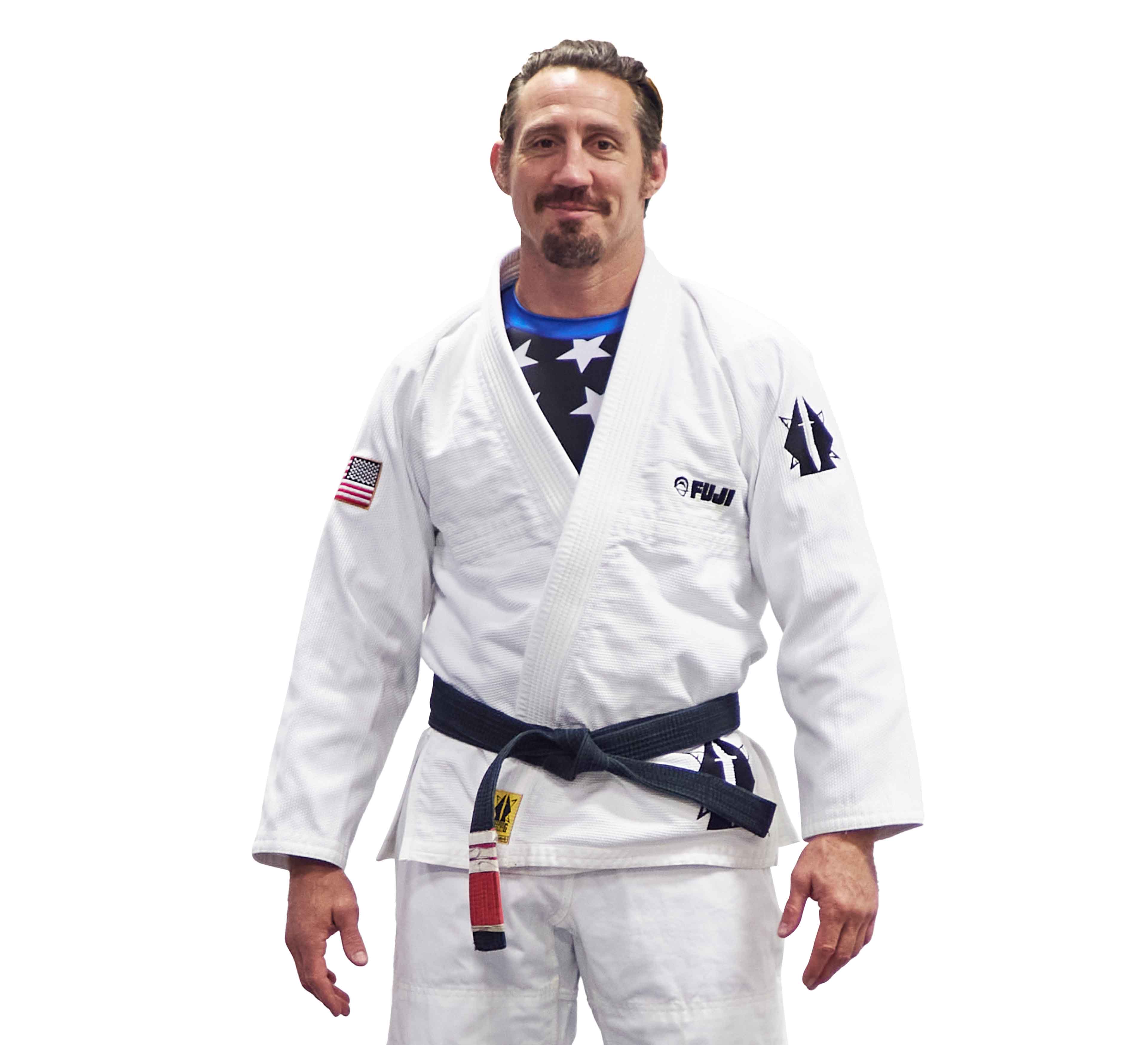 A man with short brown hair, a mustache, and a goatee stands facing forward in the Lightweight BJJ Gi Sheepdog Response Edition White with patches, including an American flag on the sleeve, and a black belt.