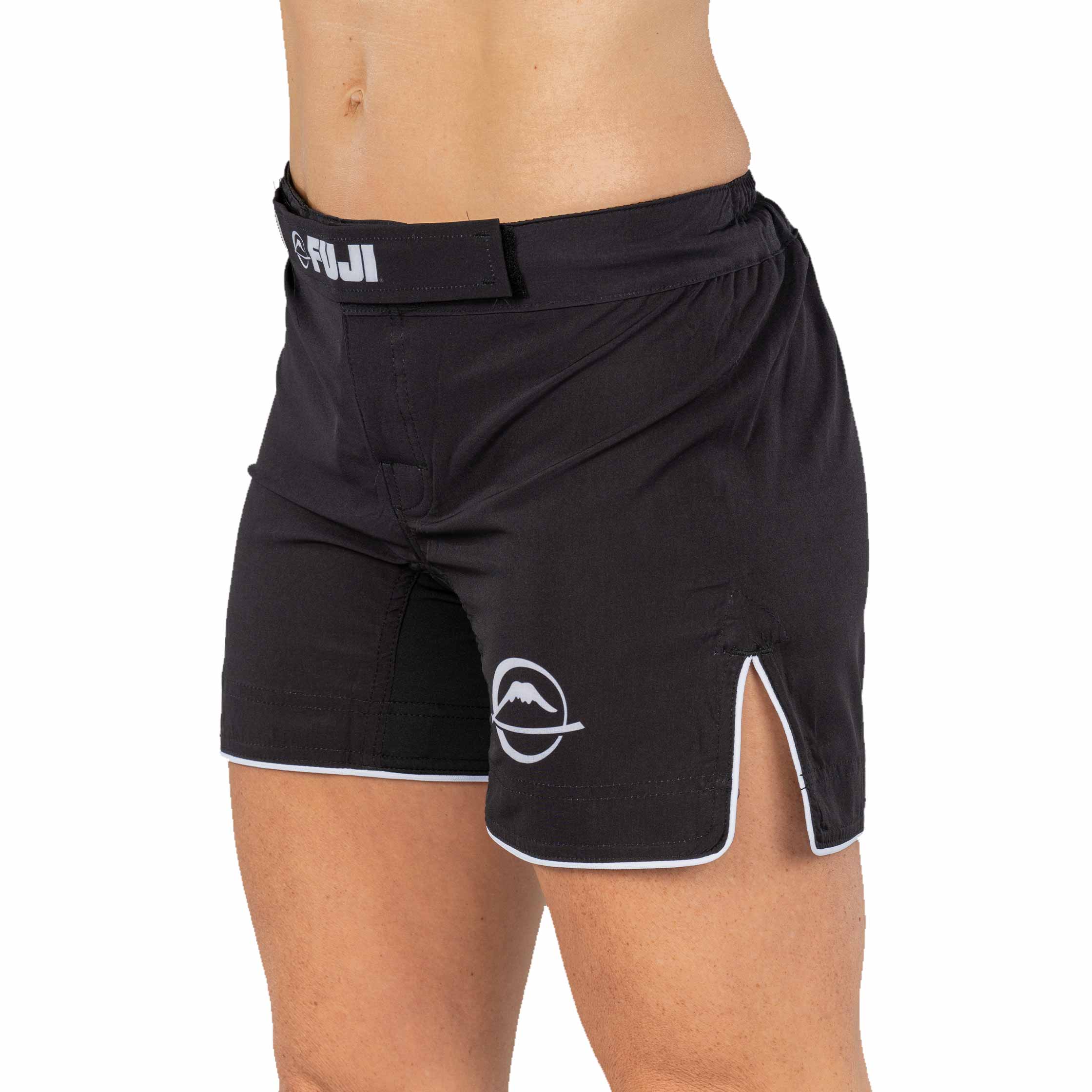 A person wearing Baseline Womens Black Grappling Shorts with white trim, a FUJI logo on the waistband, a small white mountain logo on the left leg, and side slits for mobility.