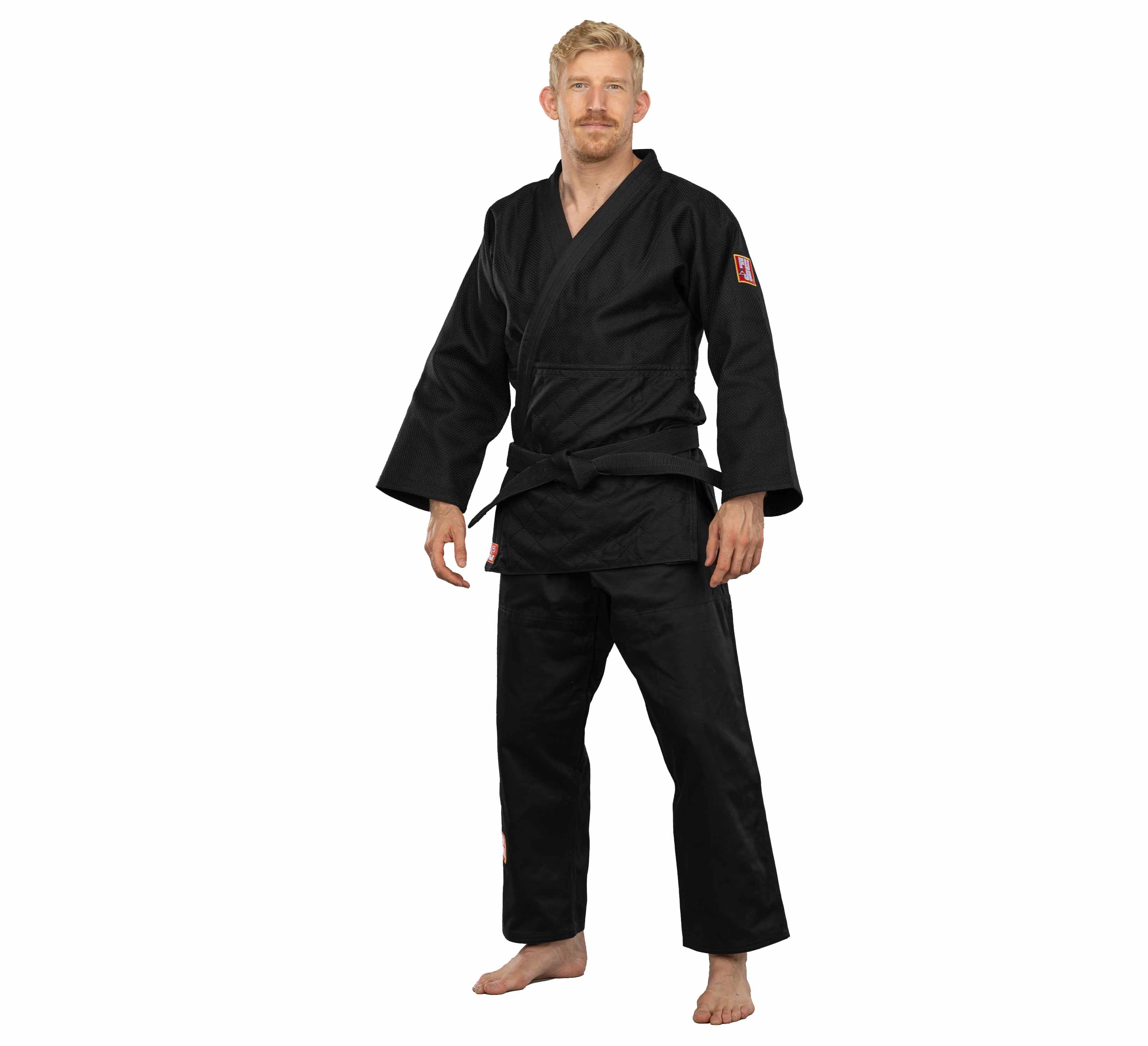 A man with short blond hair and a mustache stands barefoot against a plain white background, wearing the Euro Competition Judo Gi Black, a long-sleeve black martial arts uniform with a belt.