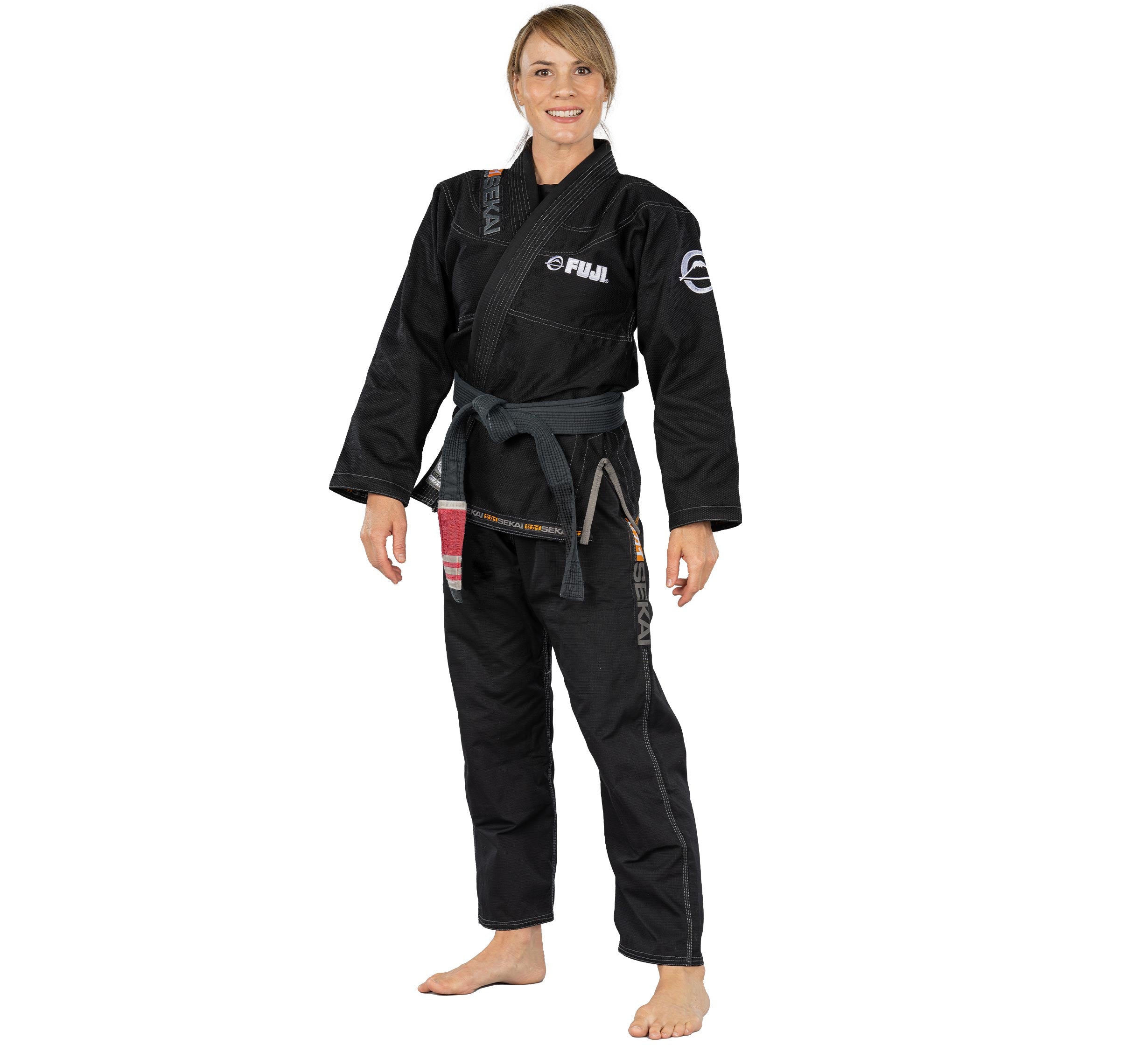 A smiling person stands barefoot against a white background, wearing the Sekai 2.0 Womens BJJ Gi Black with patches and a gray belt.
