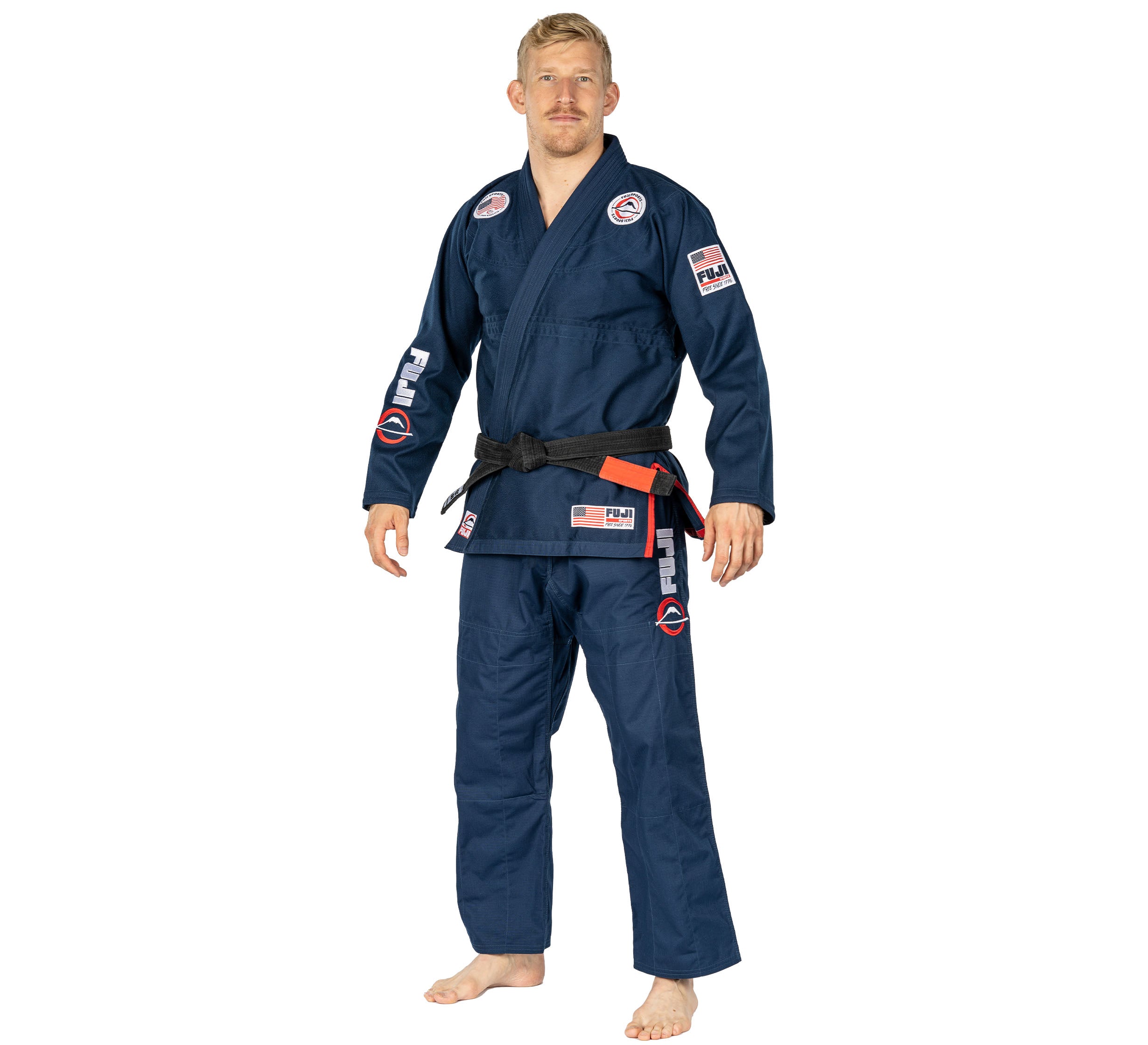 A man stands barefoot on a white background wearing the Suparaito BJJ Gi 1776 Americana Edition Navy, featuring various patches and a black belt.