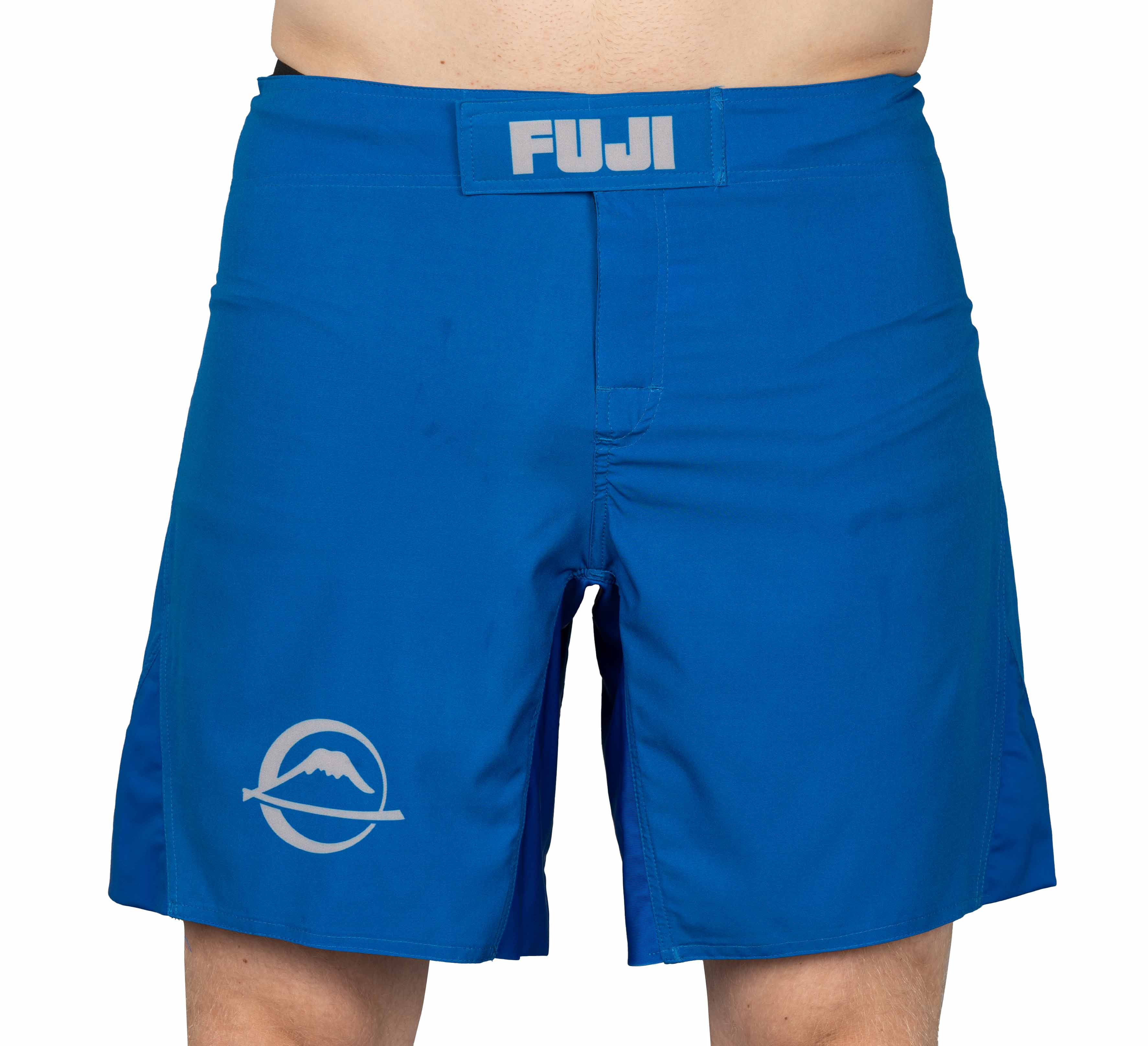 A person wearing blue Baseline Fight Shorts featuring a velcro waistband with a FUJI label, a white mountain logo on the left leg, and side slits for mobility.