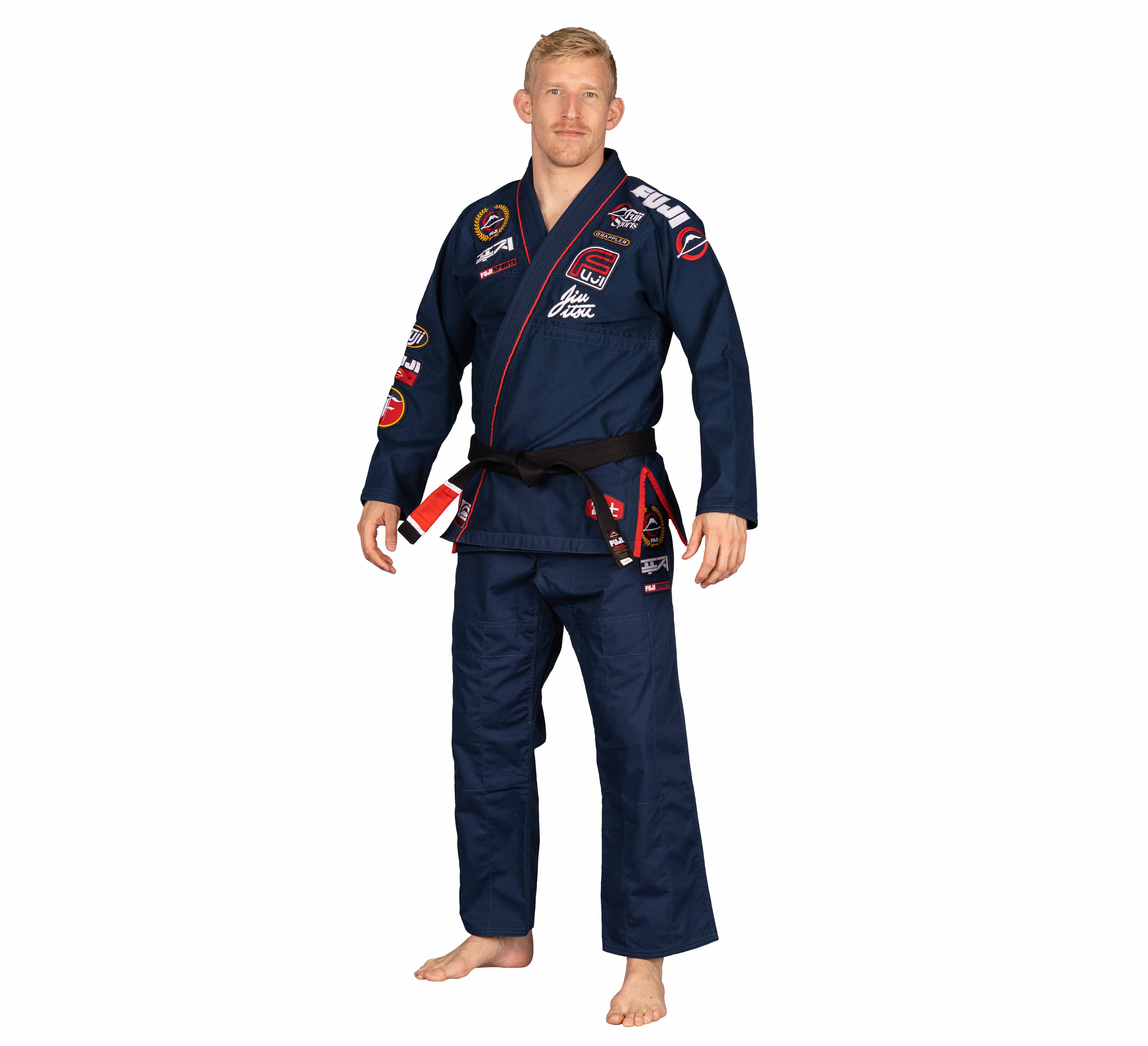 A man wearing a Suparaito BJJ Gi XTR Edition Navy with multiple patches and a black belt stands barefoot on a white background.