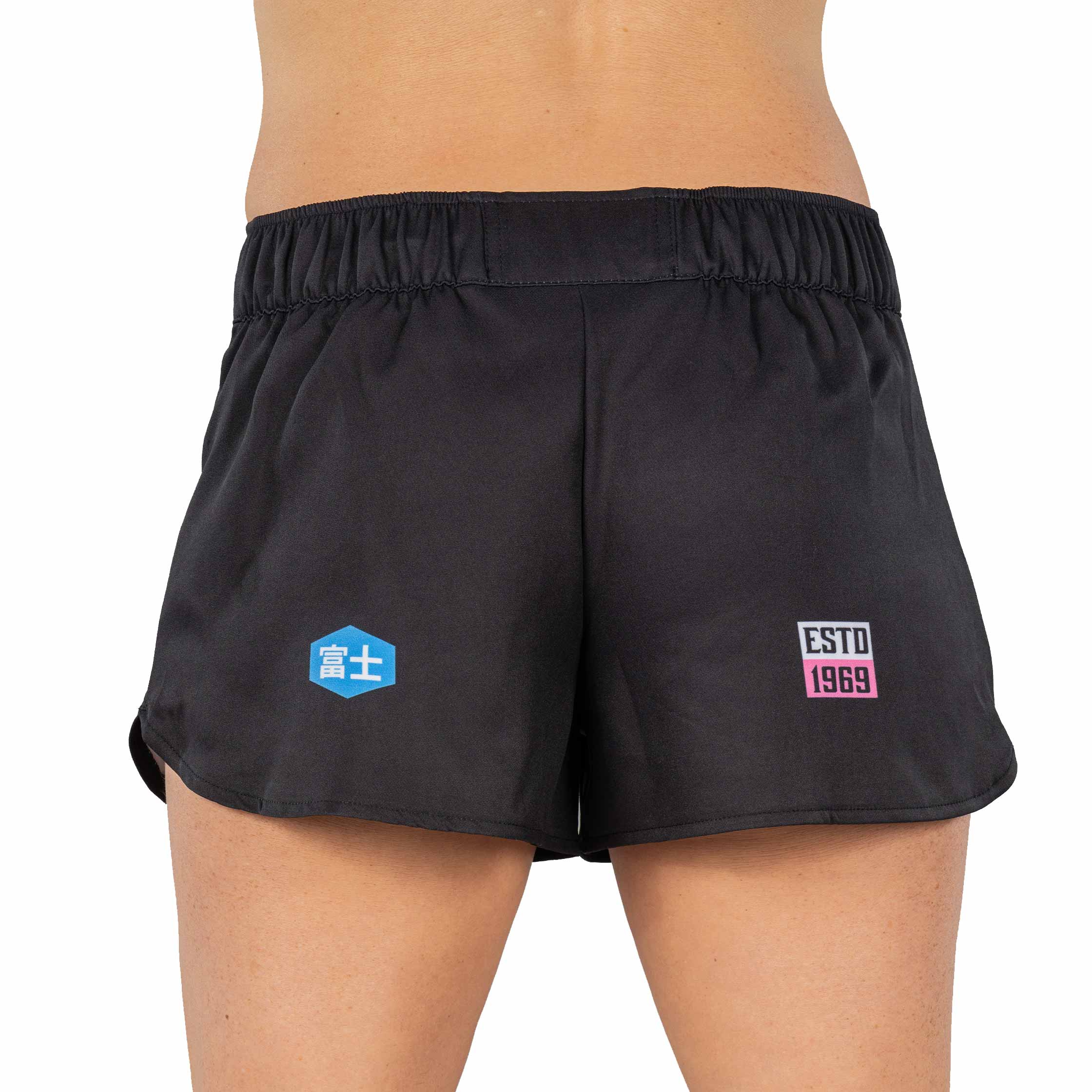 A person wearing XTR Extreme Womens Grappling Fight Shorts Pink, viewed from the back. The shorts feature a blue hexagonal logo on the left side and a rectangular “ESTD 1969” patch on the right.