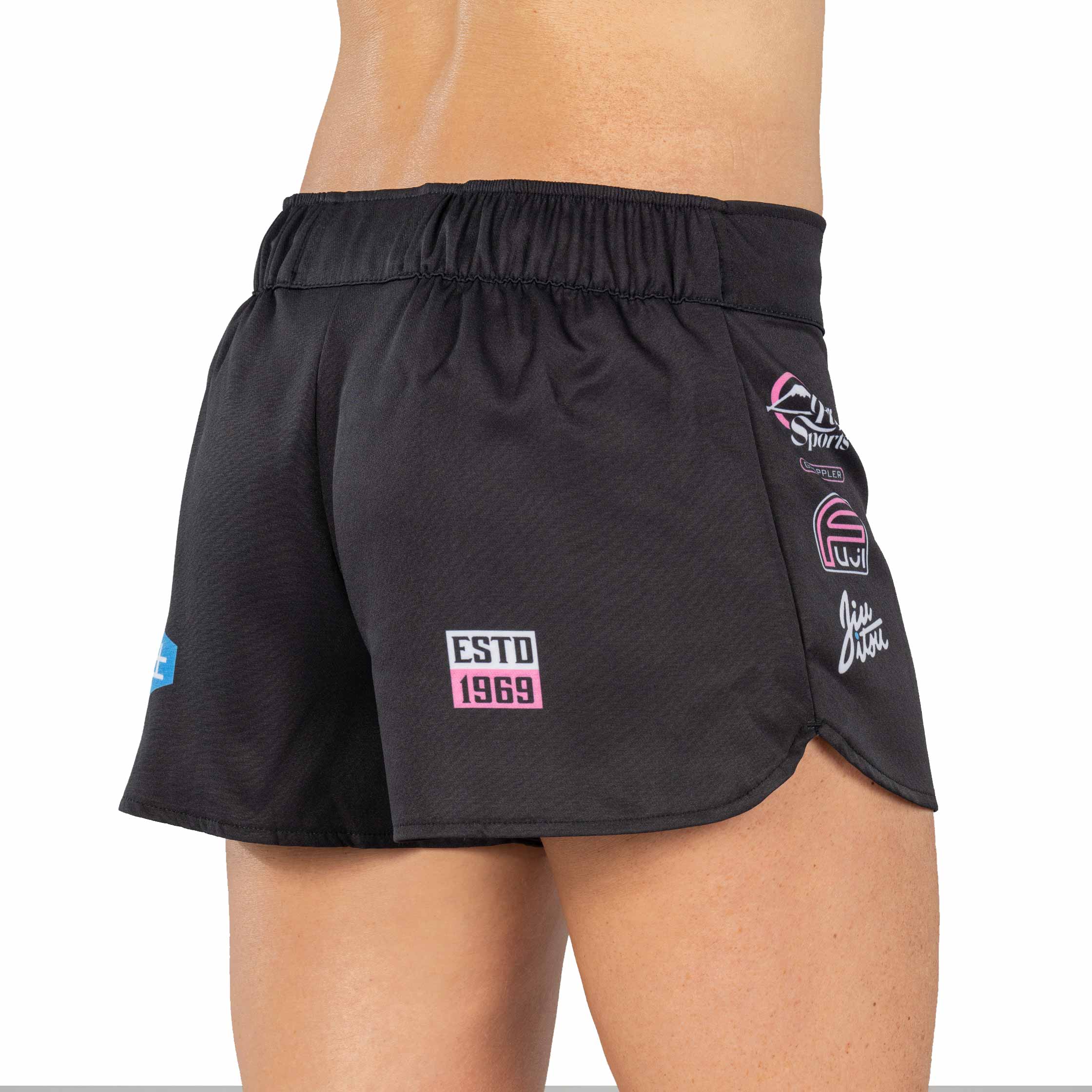 A person wearing XTR Extreme Womens Grappling Fight Shorts Pink, featuring black fabric with colorful patches and ESTD 1969 on the back. The image displays the waist and upper legs from behind.