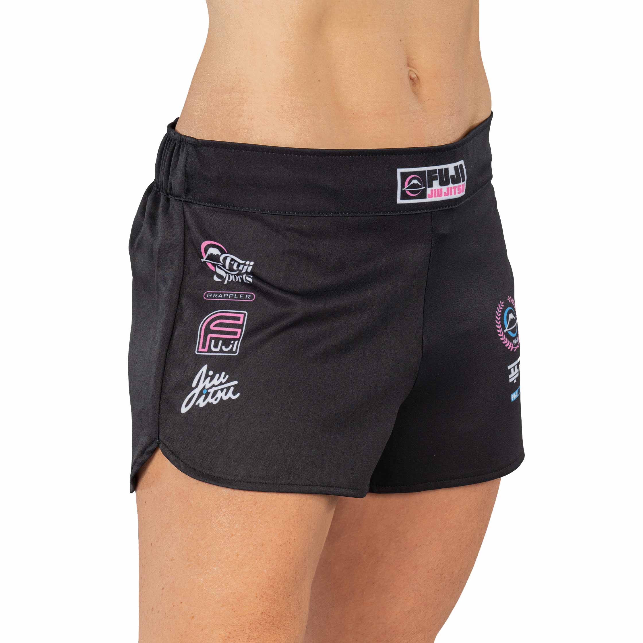 A person is wearing XTR Extreme Womens Grappling Fight Shorts Pink, featuring various martial arts and brand logos, including FUJI and Jiu Jitsu, printed on the front. Their upper thighs and lower torso are visible.