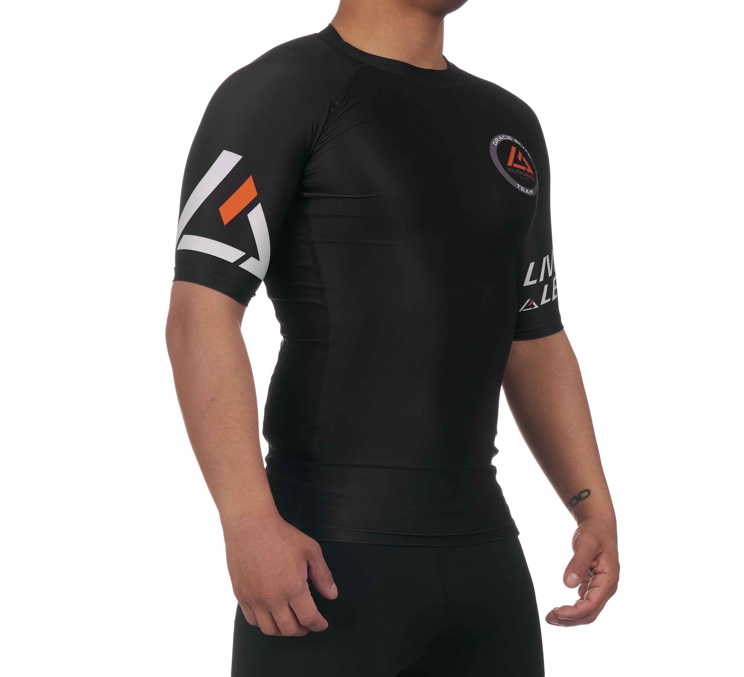 A person wears the Rilion Gracie Ranked Short Sleeve Rashguard in black, featuring white and orange accents with logos on the sleeve and chest, standing against a white background.