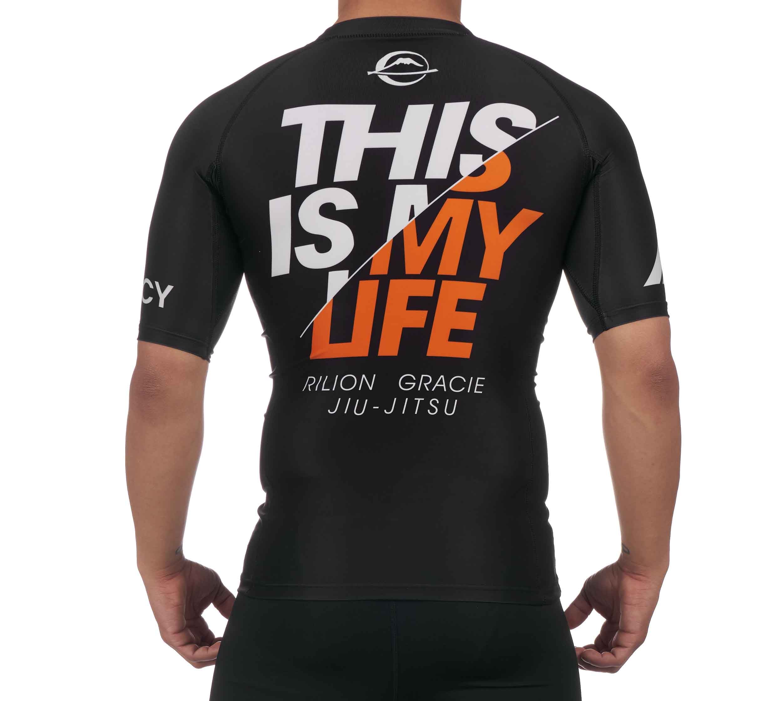 A person wearing the Rilion Gracie Ranked Short Sleeve Rashguard Black, featuring bold white and orange "THIS IS MY LIFE" text on the back with "RILION GRACIE JIU-JITSU" in smaller letters beneath.