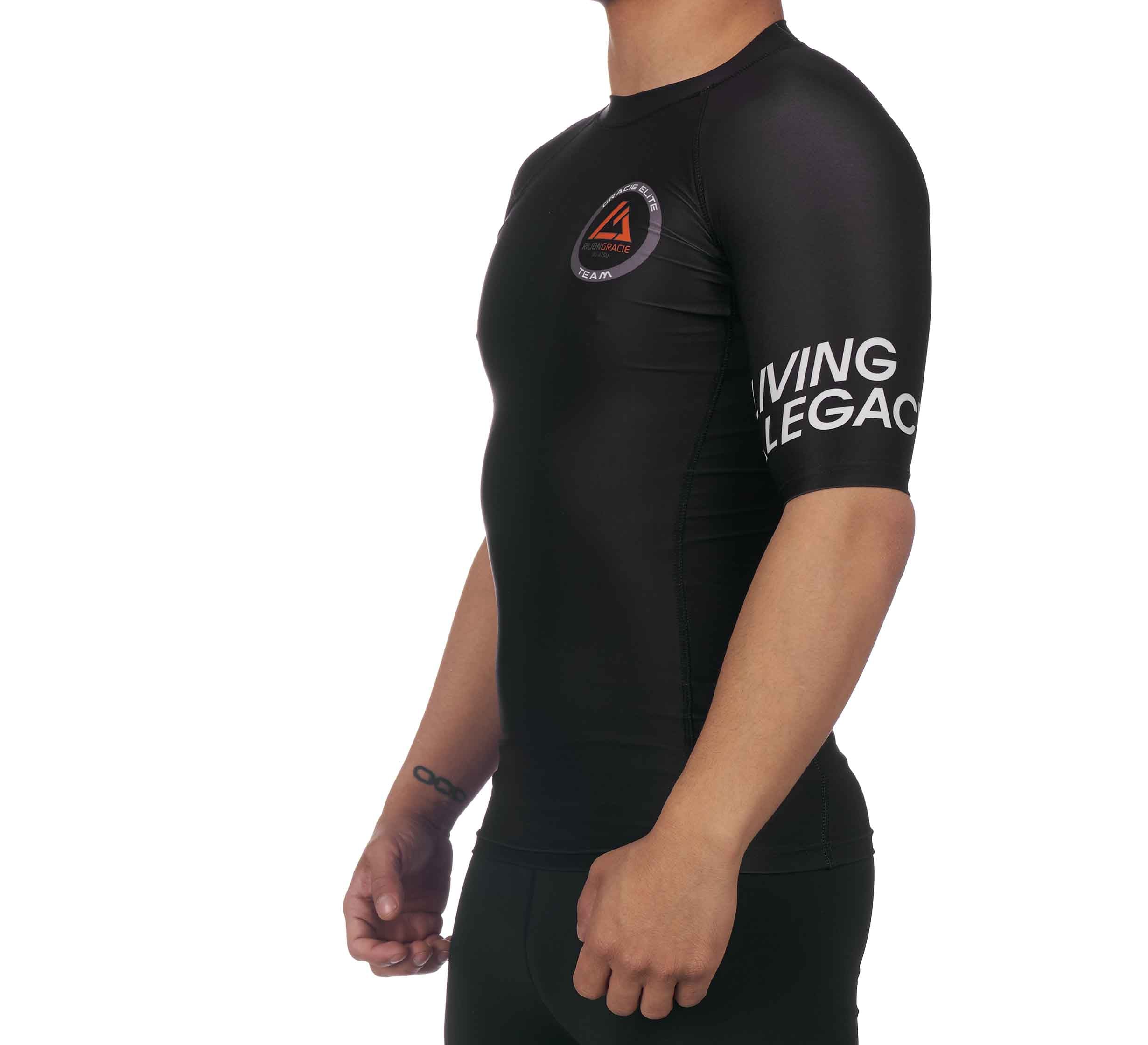 Someone wearing the Rilion Gracie Ranked Short Sleeve Rashguard Black, featuring "LIVING LEGACY" on the sleeve and a circular logo on the chest, stands against a white background.