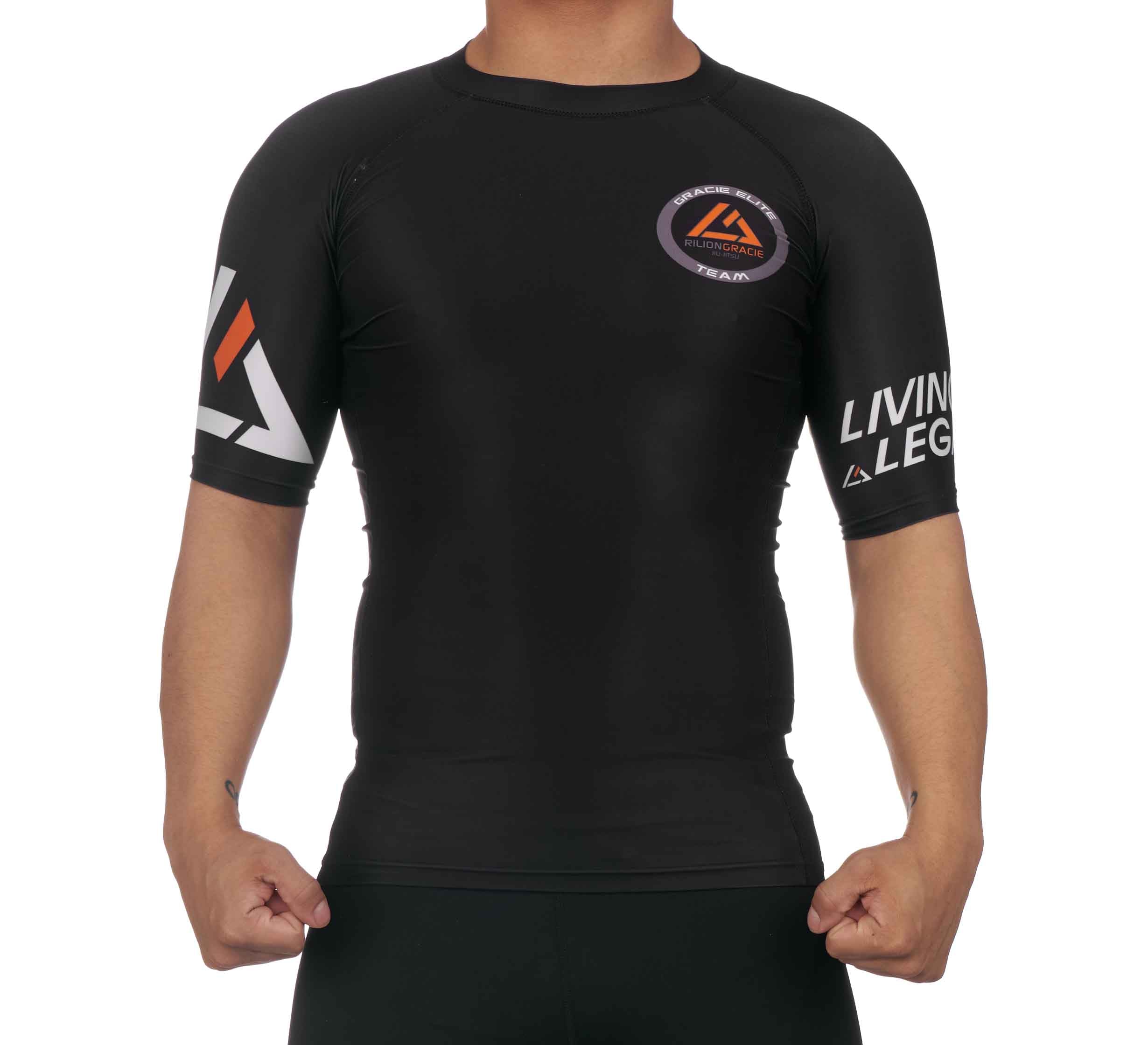 A person wearing the Rilion Gracie Ranked Short Sleeve Rashguard Black, featuring logos and text on the chest and sleeves, stands with fists clenched at their sides, facing the camera.