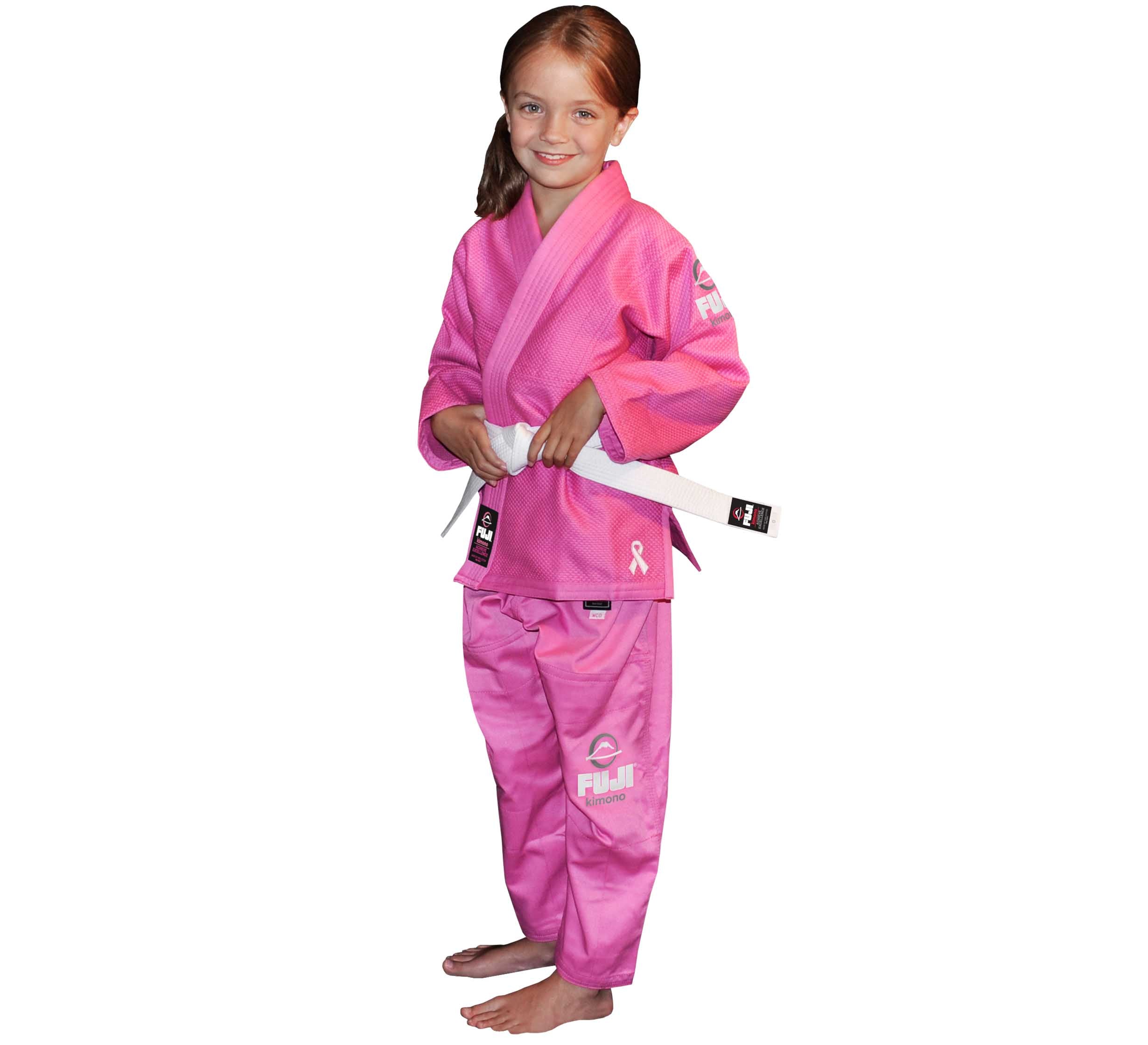 A young girl smiles at the camera, barefoot and wearing the Pink All Around Kids BJJ Gi with a white belt. The gi features logo details and a pink ribbon symbol on the jacket.