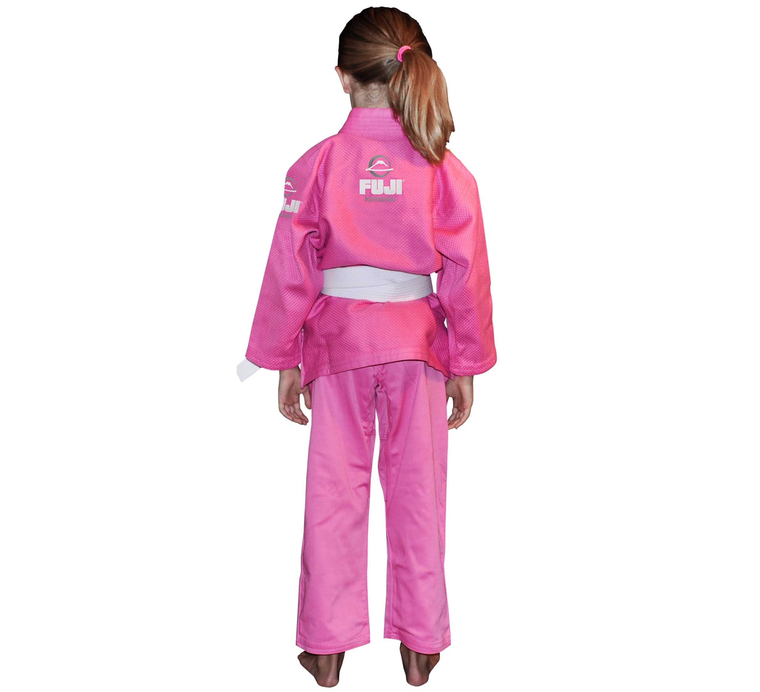 A young girl with brown hair in a ponytail stands barefoot, facing away, wearing the Pink All Around Kids BJJ Gi with a white belt and FUJI logos and text on the back.