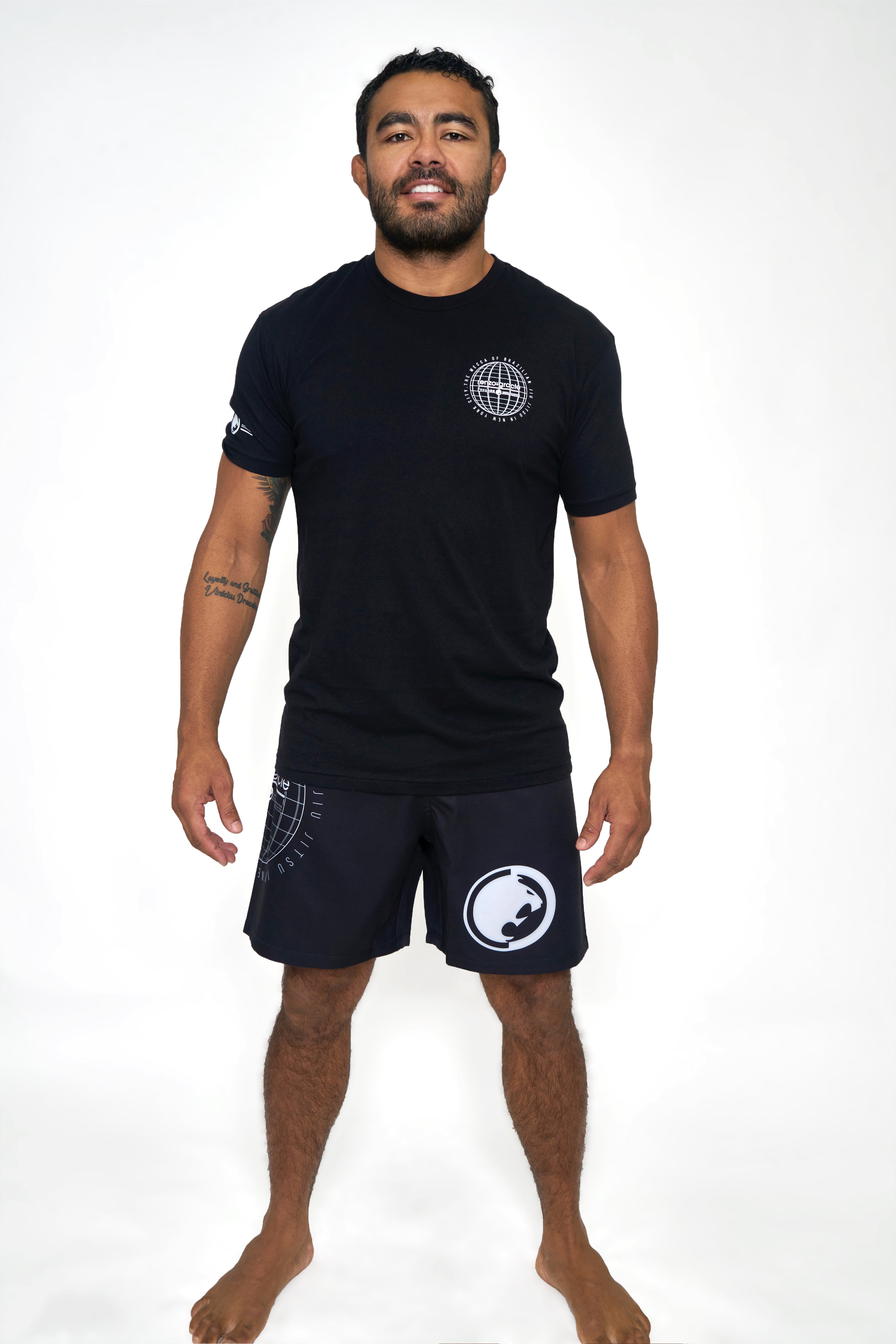 A man with short dark hair, a beard, and tattoos on his left forearm stands barefoot on a white background, smiling. He wears the Renzo Gracie Jiu Jitsu Mecca T-Shirt in black and matching black athletic shorts with printed logos.