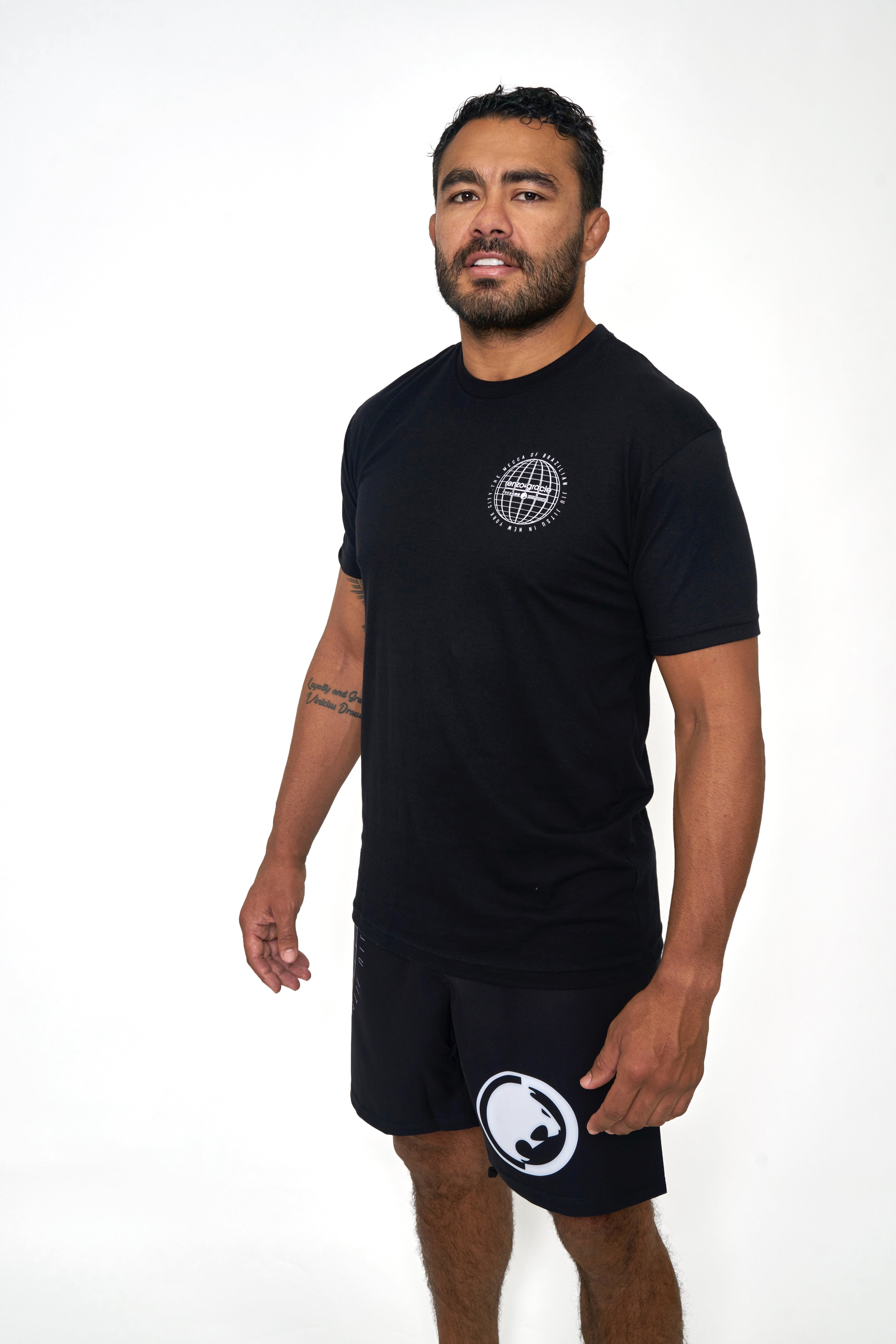 A man with short dark hair and a beard stands against a white background, wearing the Renzo Gracie Jiu Jitsu Mecca T-Shirt in black with a small white globe logo, black athletic shorts with a white circular logo, and showing off his tattoo as he smiles slightly.