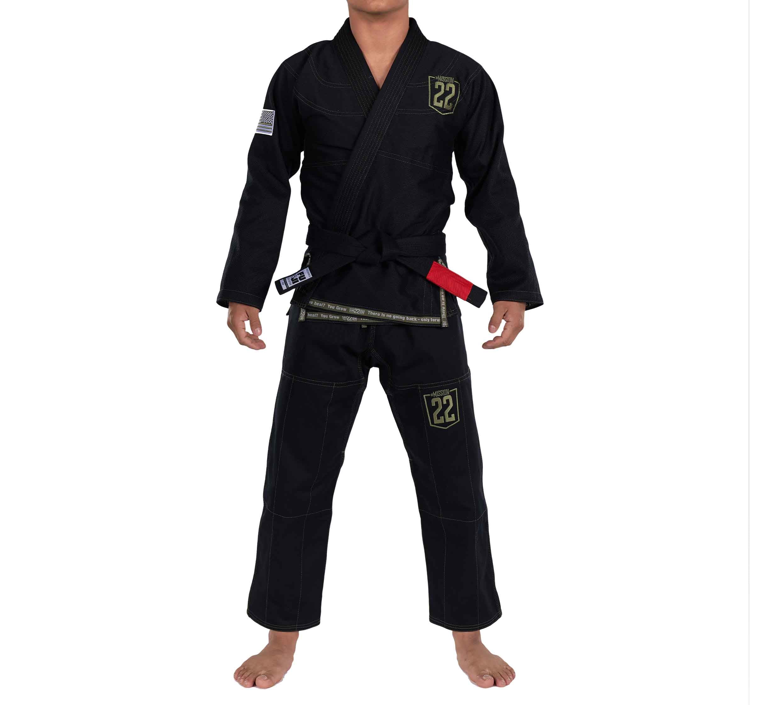 A person wearing the Mission 22 Black Gi with a black belt stands barefoot against a white background. The gi features yellow patches with the number 22 on the chest and left leg, and an American flag patch on the right arm.